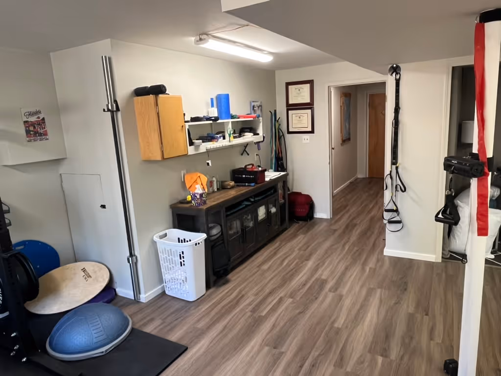 Home gym room with wood flooring, wall-mounted TRX suspension straps, exercise equipment, storage cabinet, and a laundry basket. cadence-performance