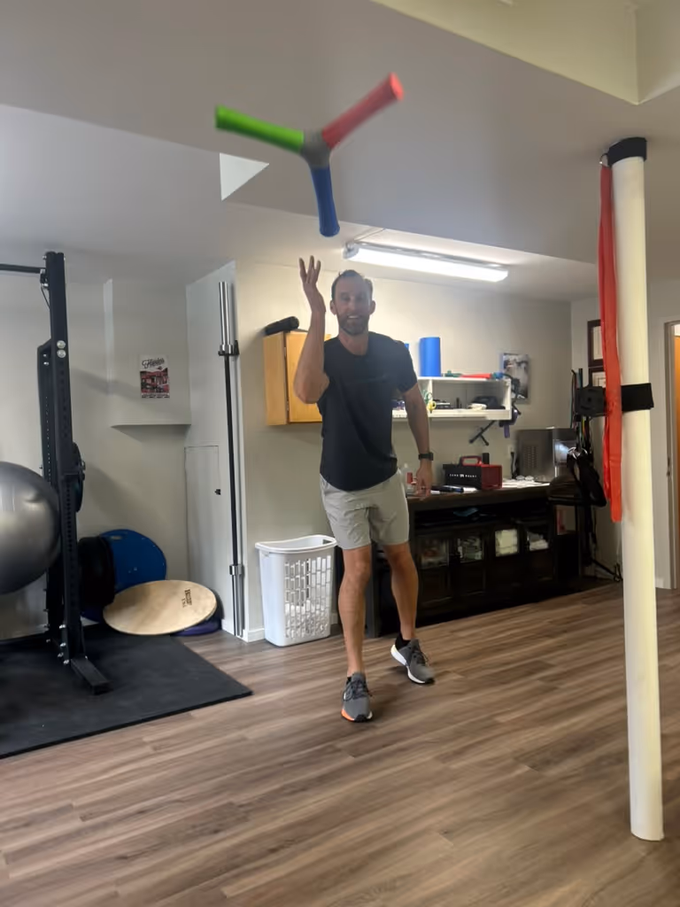 Man indoors juggling or tossing a three-pronged red, green, and blue object, standing on wood-patterned floor in a room with exercise equipment. cadence-performance