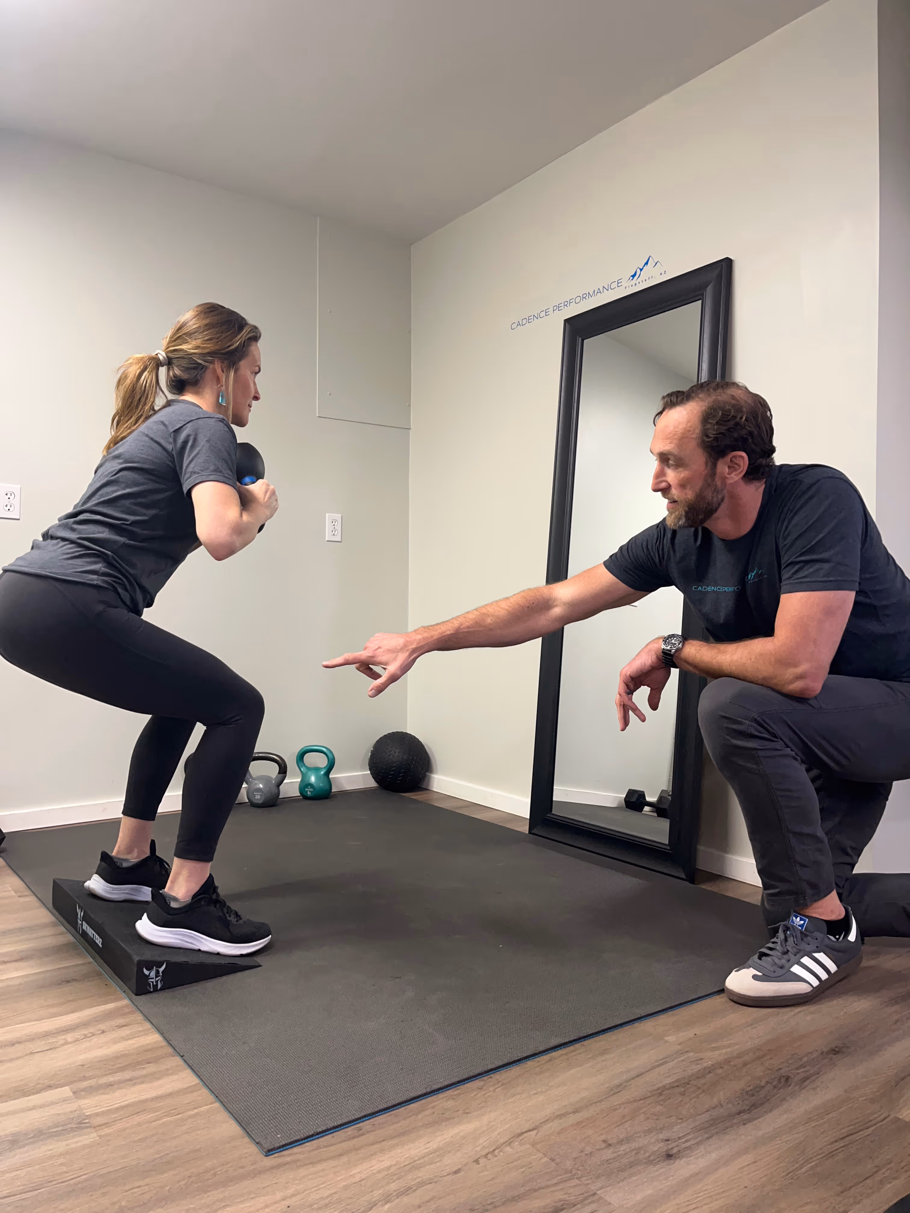 Woman performing a kettlebell squat on a Balance Trainer platform while a trainer kneels and points, offering guidance in a fitness studio.