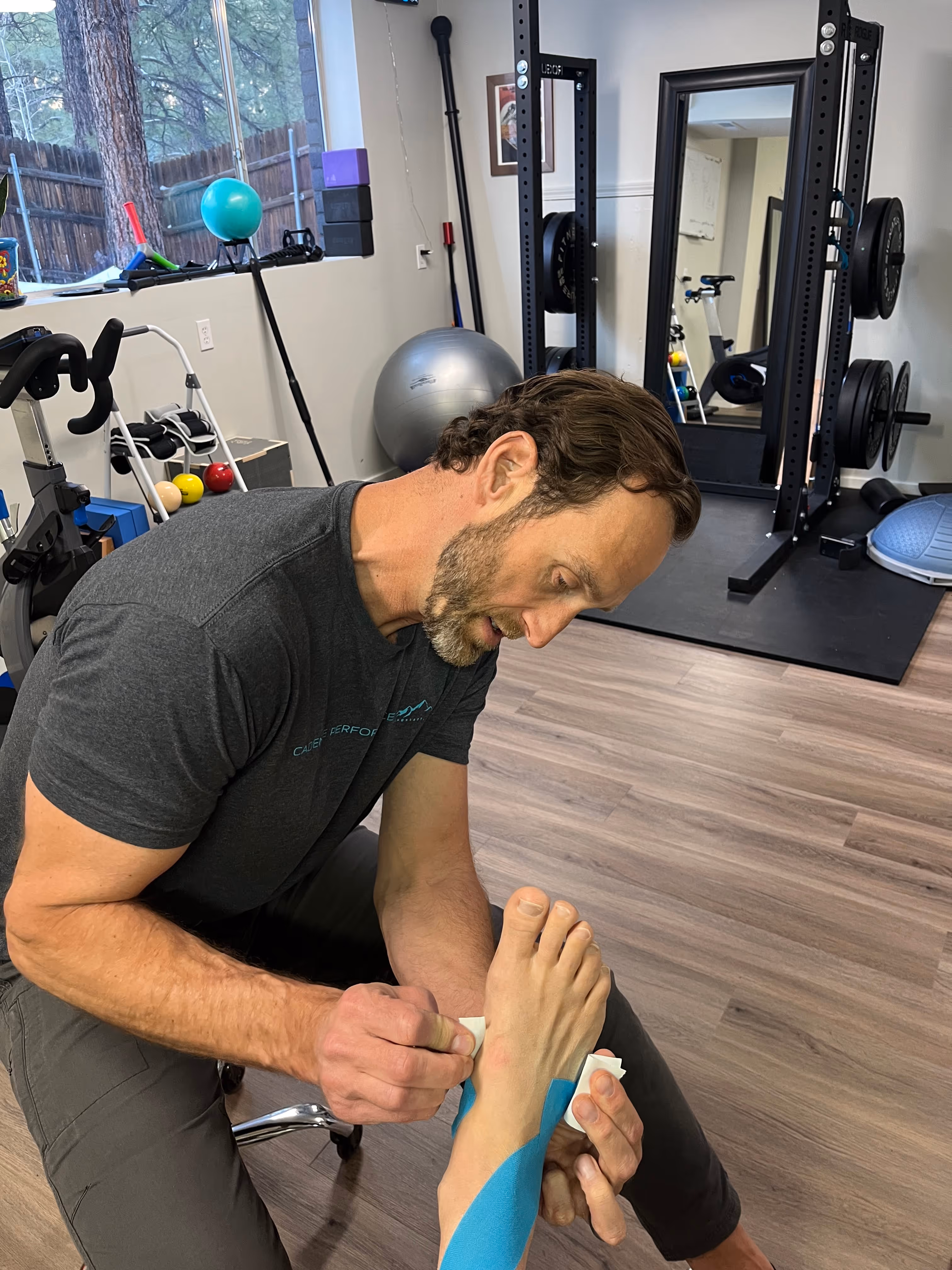 Man applying blue kinesiology tape to a person's foot in a home gym setting.