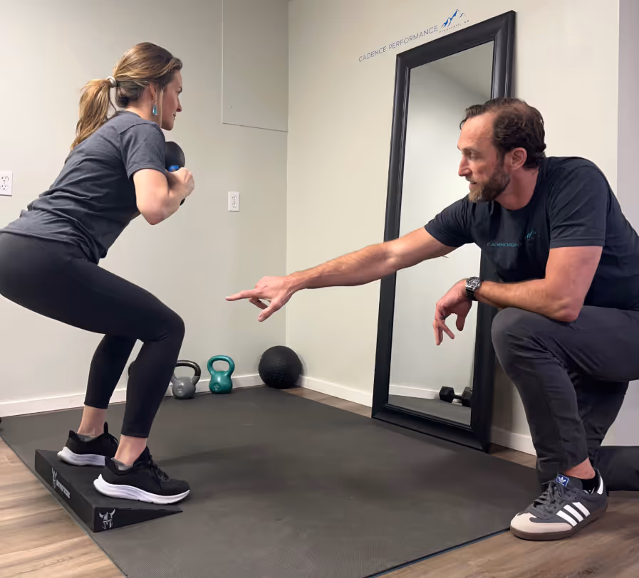 Woman performing weighted squats on a platform while a coach points and guides her posture in a fitness studio.