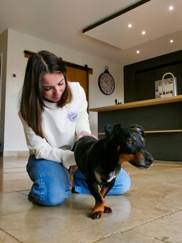 Une femme en sweat blanc caresse un petit chien noir et marron sur un sol carrelé intérieur.