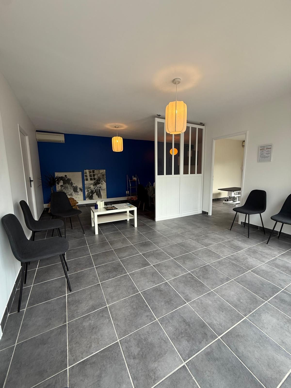 Salle d'attente moderne avec chaises noires, table blanche, murs blanc et bleu, et lampes suspendues.