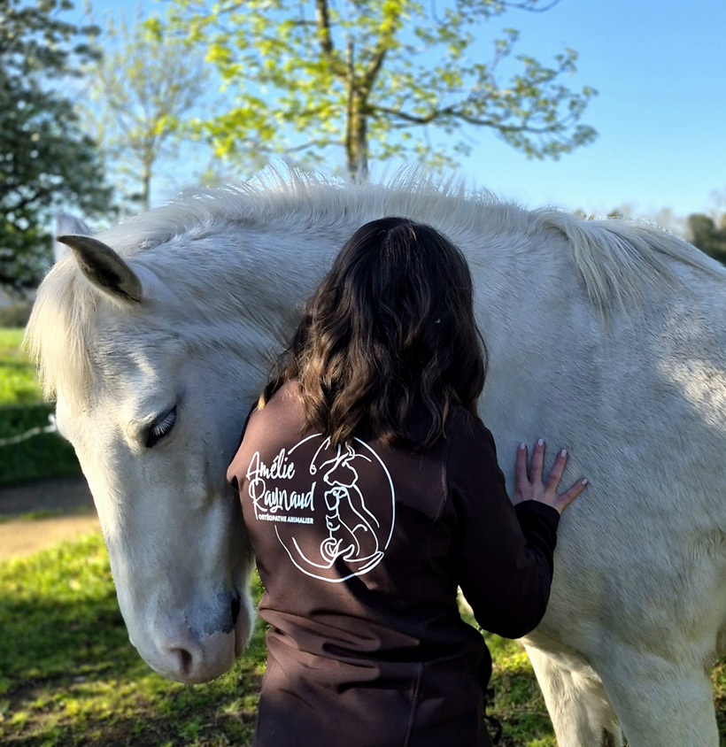 Une femme caresse un cheval blanc dans un champ sous un ciel bleu clair.