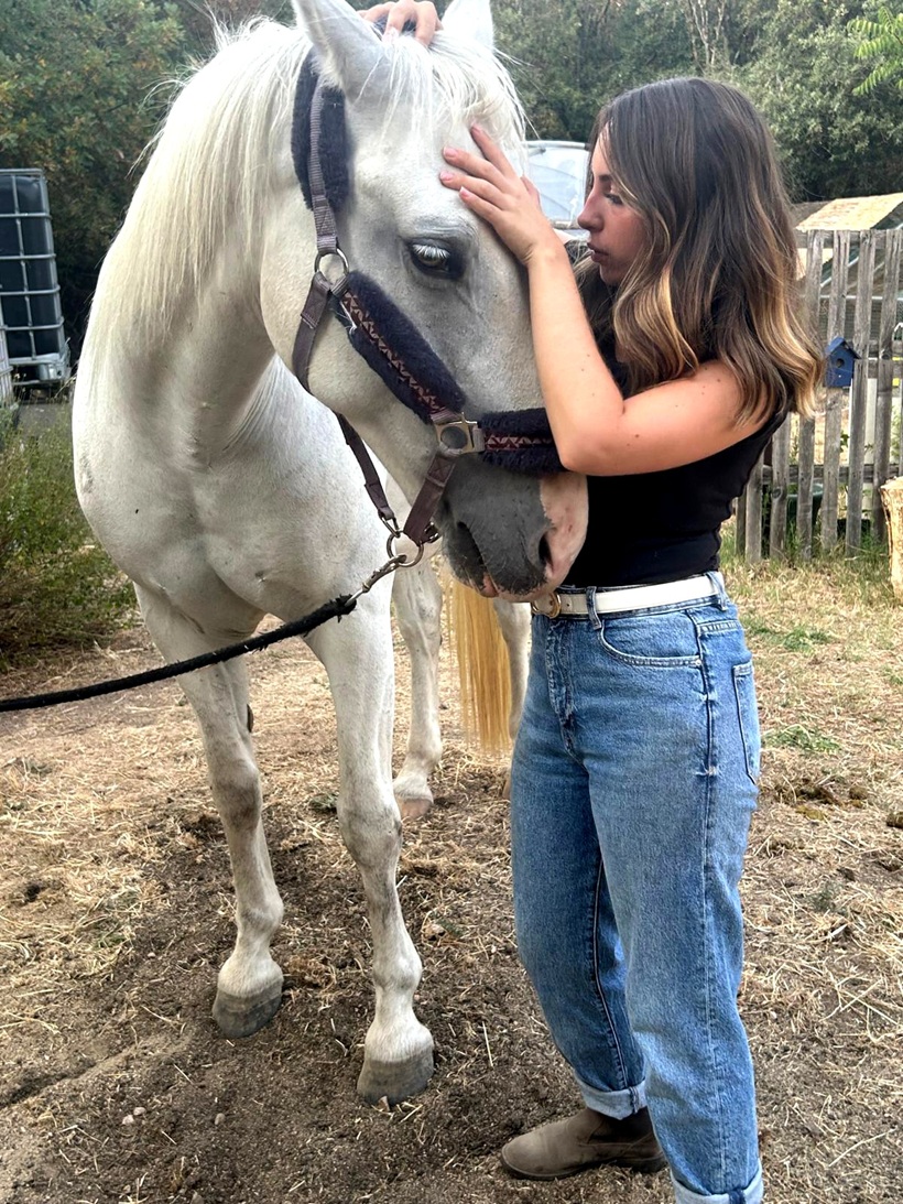 Jeune femme caressant la tête d’un cheval blanc dans un enclos extérieur.