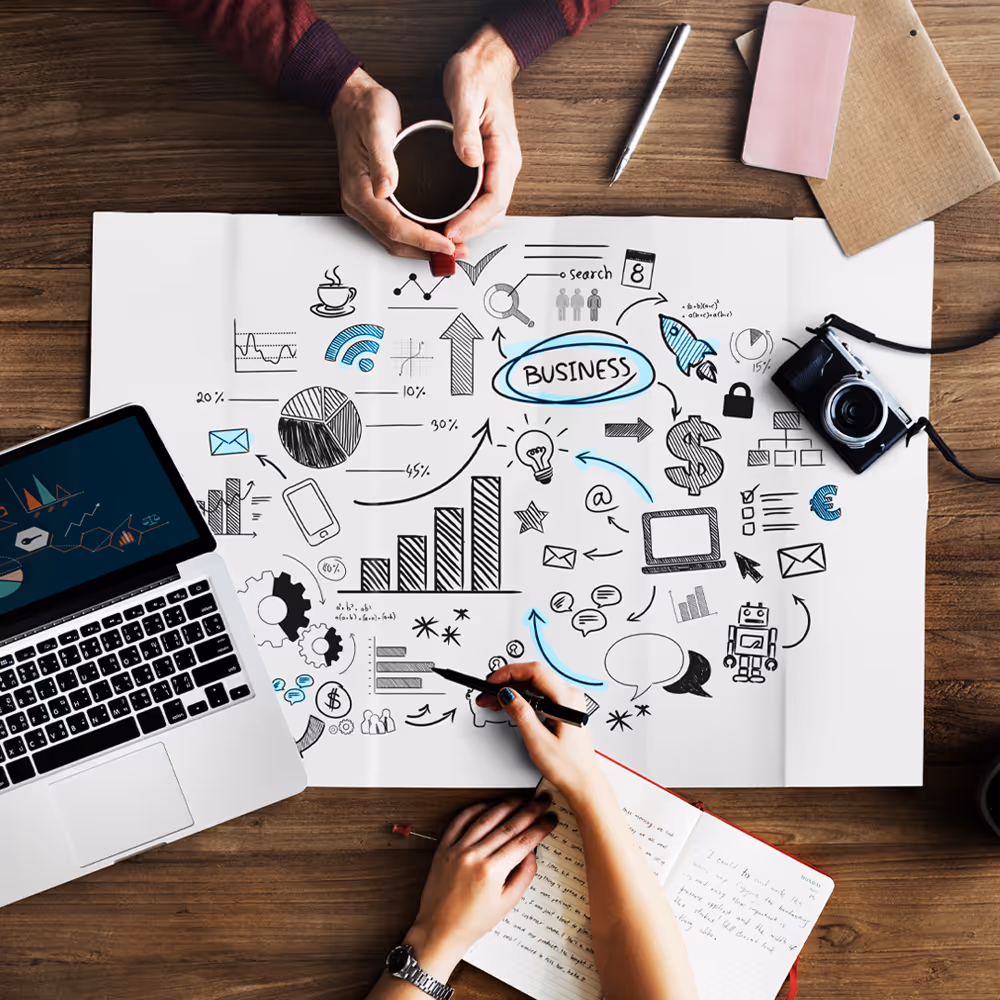 Overhead view of hands holding a coffee cup and writing in a notebook next to a laptop and paper with business and technology sketches on a wooden table.