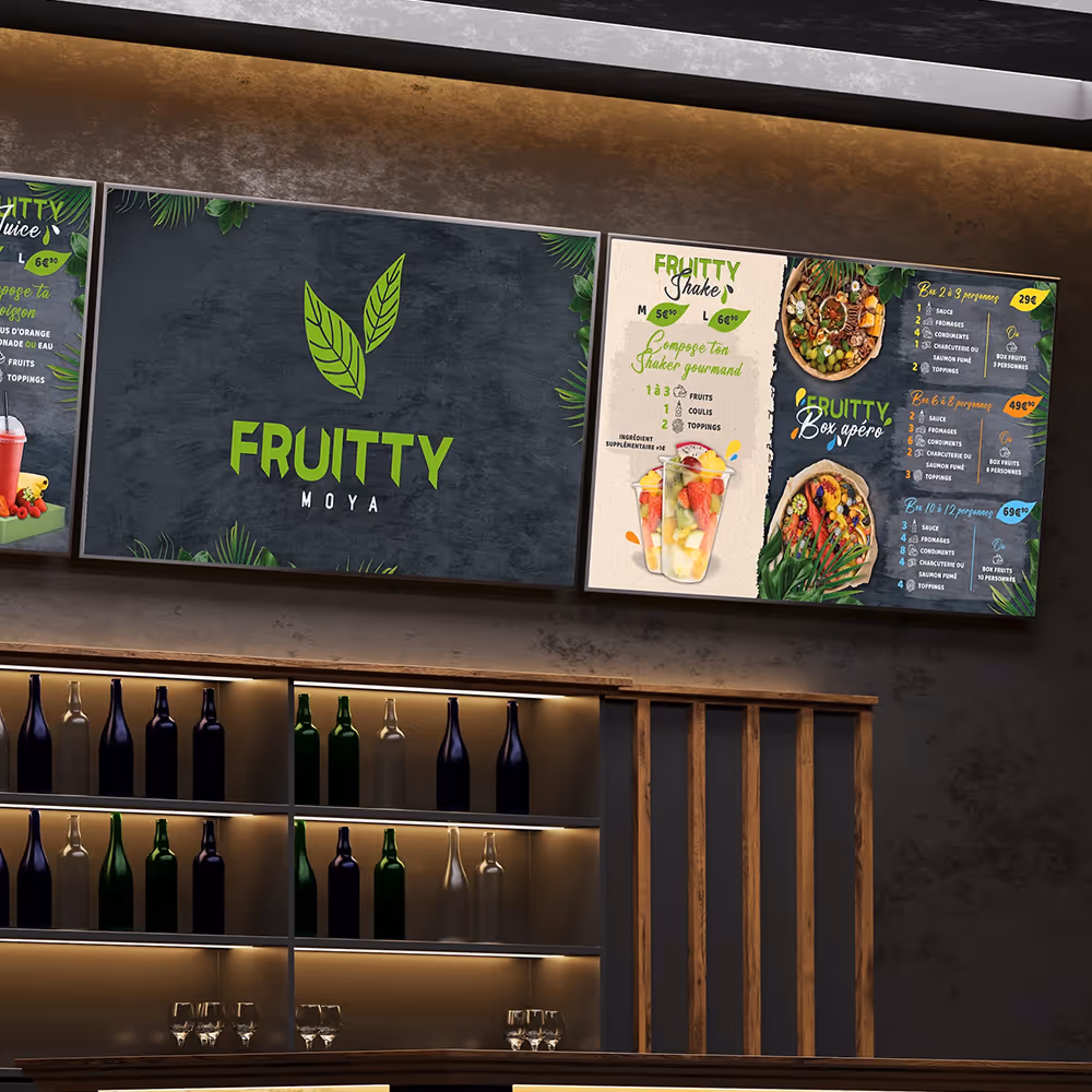 Wall-mounted digital menu boards above a bar shelf displaying Fruitty MOYA branding, fruit shake options, and box apéro packages.