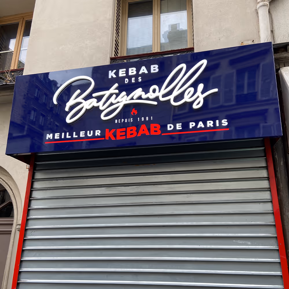 Blue sign above a closed metal shutter reading 'Kebab des Batignolles, Depuis 1991, Meilleur Kebab de Paris' with red and white text.