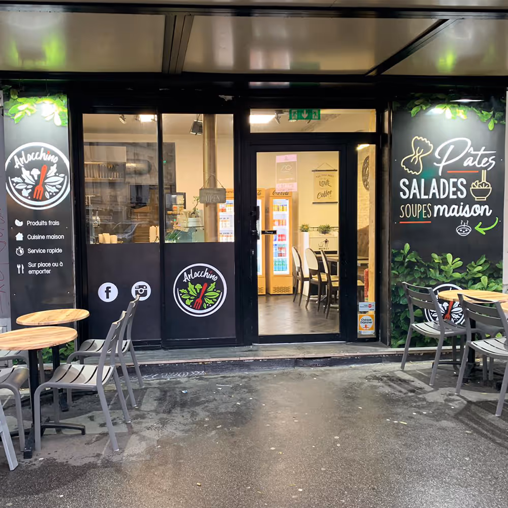 Front entrance of a small restaurant with outdoor tables and chairs, black doors with logos and signs advertising fresh products, homemade cuisine, and pasta salads.