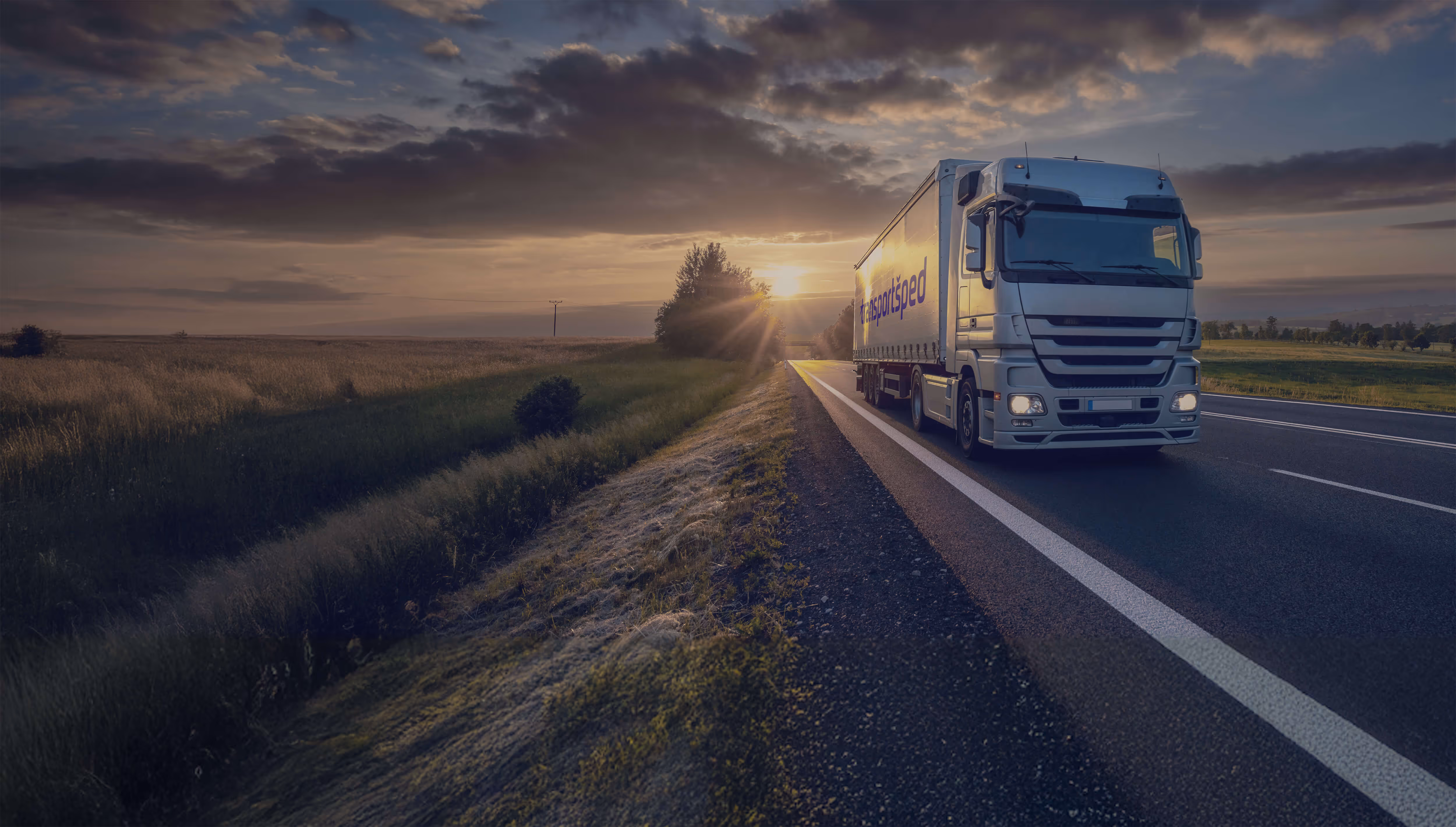 White transport truck driving on a highway at sunset with fields and trees on the side.