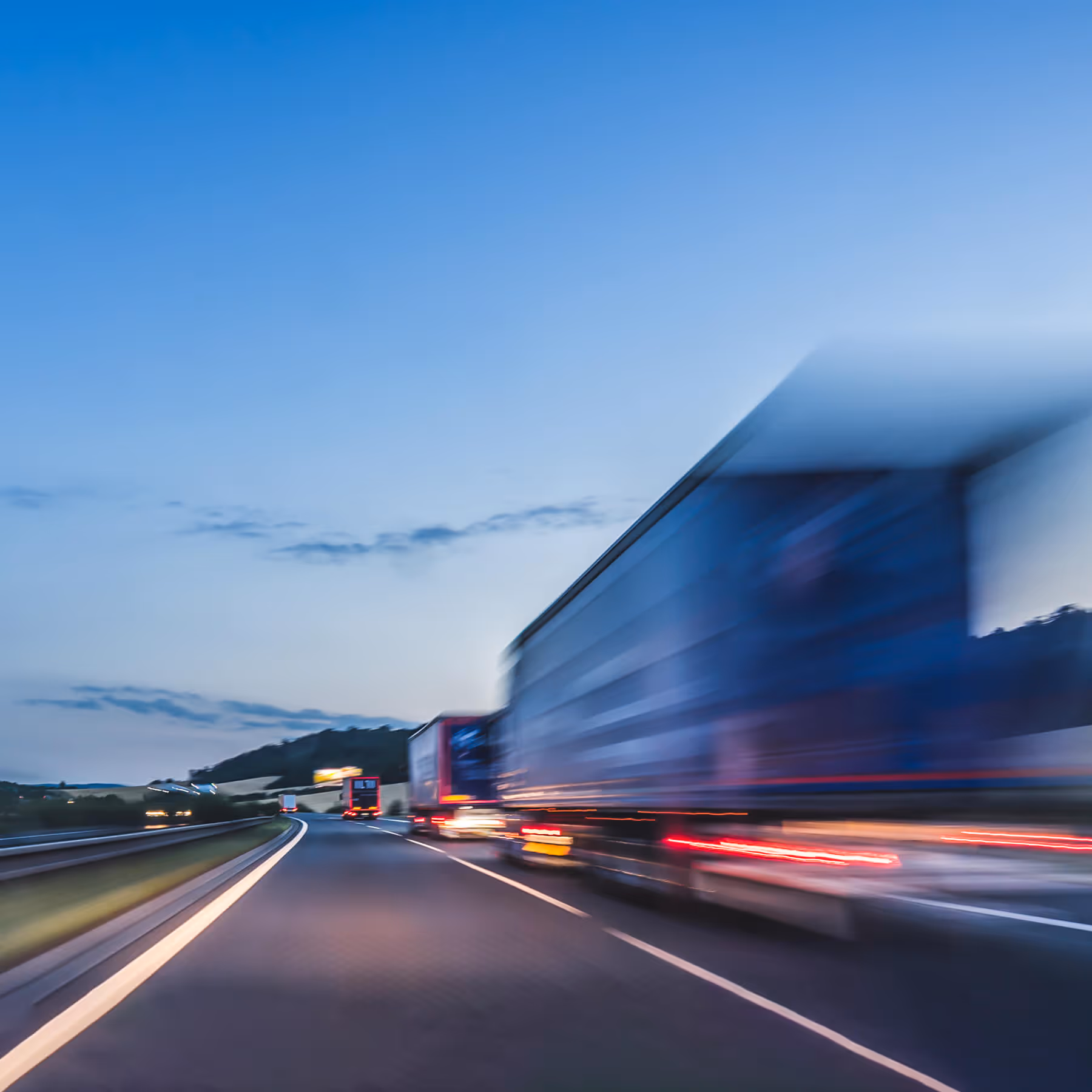Blurred motion of several large trucks driving along a highway at dusk with a clear blue sky.