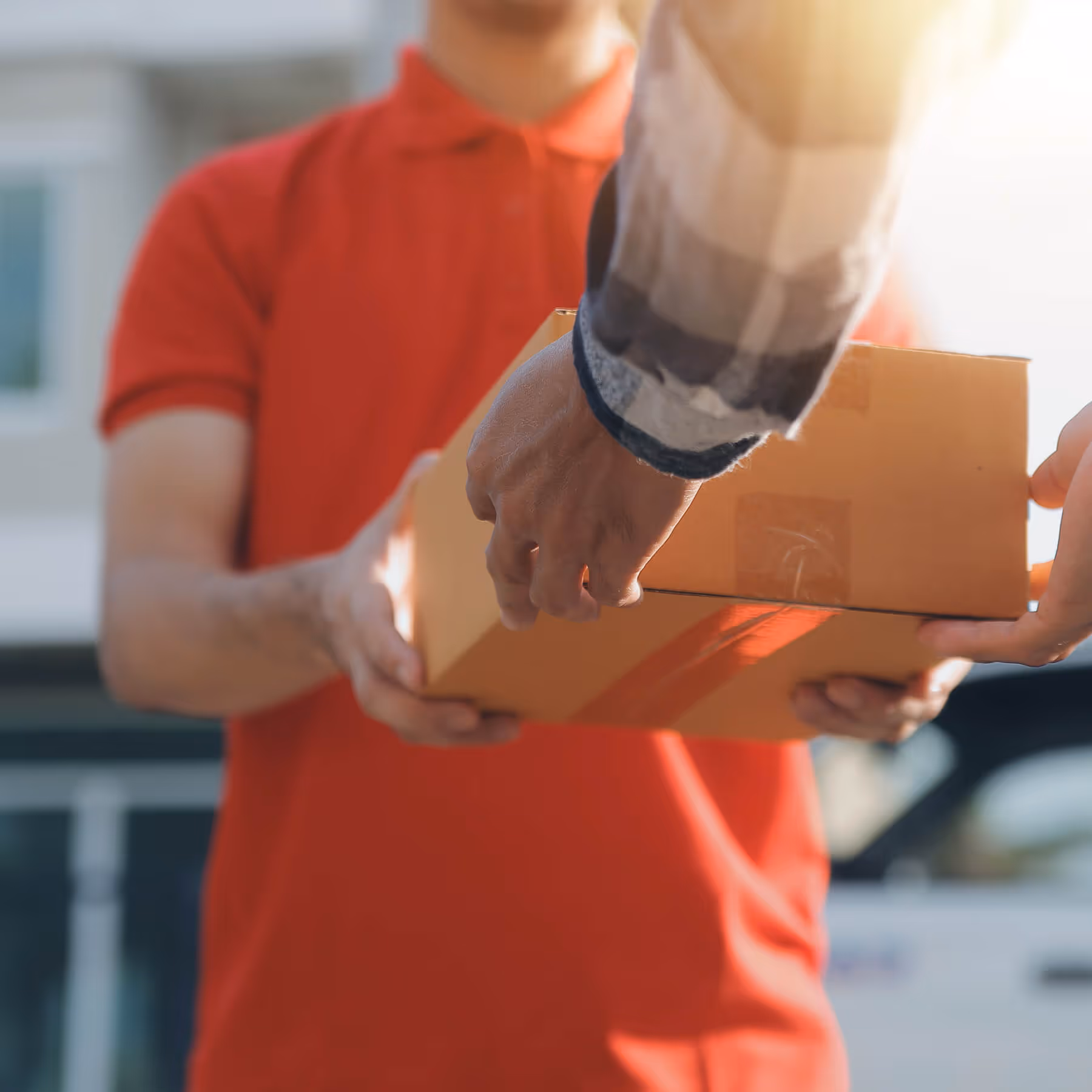 Person in red shirt handing a cardboard package to another person outdoors in sunlight.