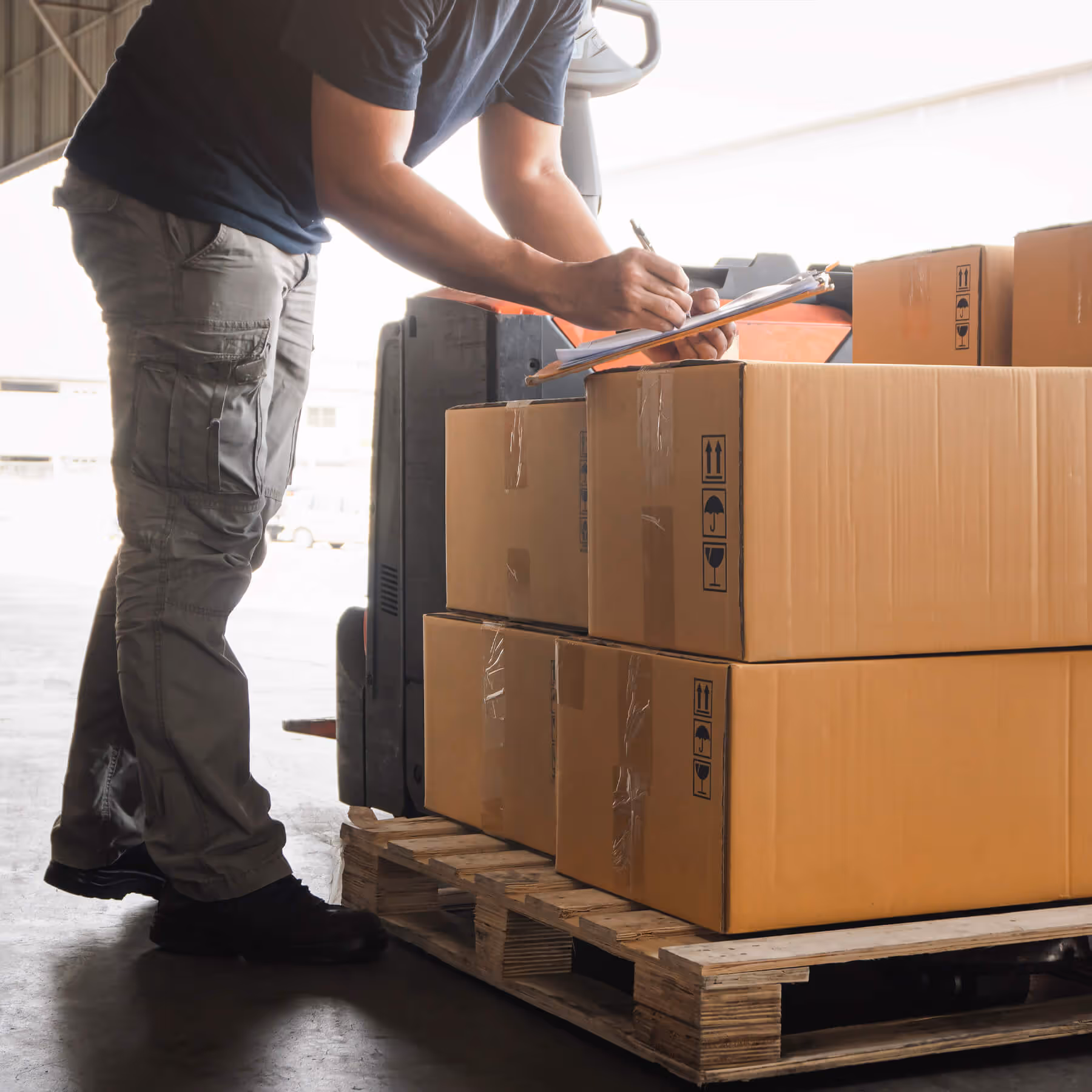 Worker checking and marking inventory on a clipboard beside a pallet stacked with cardboard boxes in a warehouse.