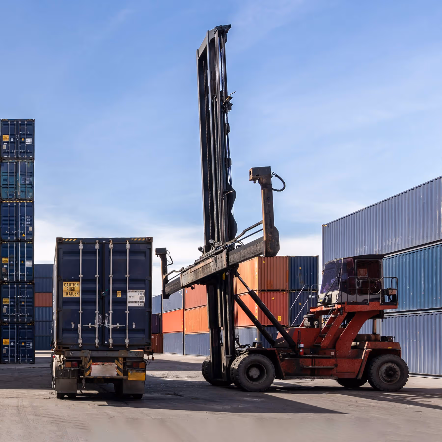 Large forklift lifting a shipping container next to a truck trailer and stacked colorful shipping containers under a clear blue sky.