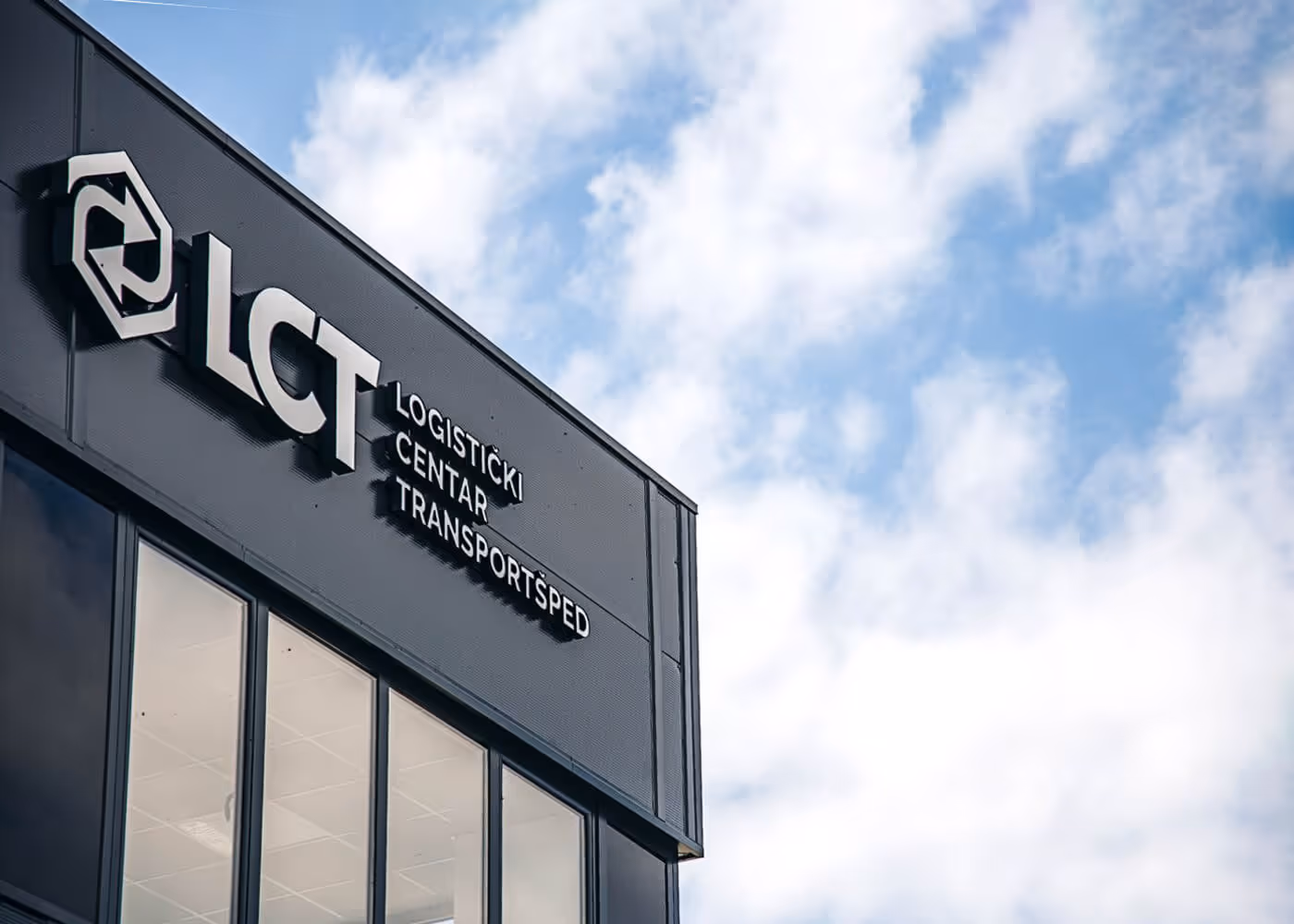 Exterior of a modern building with the sign 'LCT Logistički Centar Transportsped' against a partly cloudy sky.