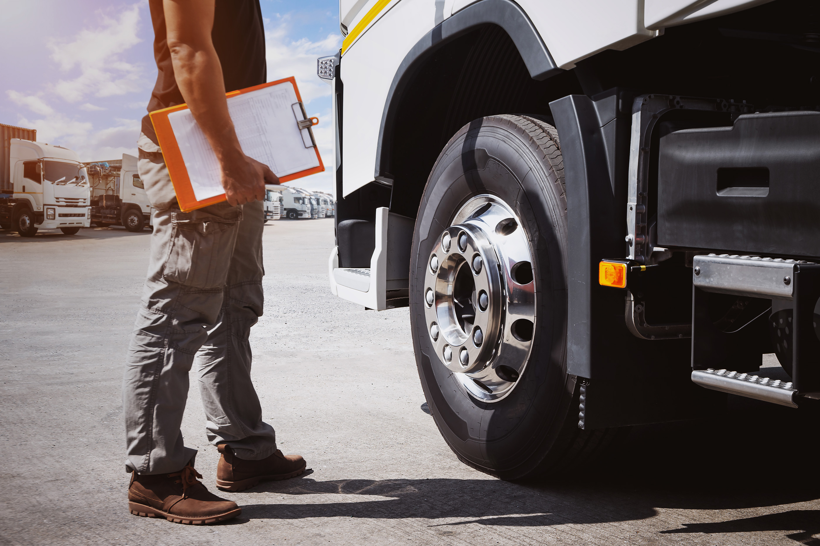 Man holding clipboard inspecting the front tire of a white truck in a truck yard.