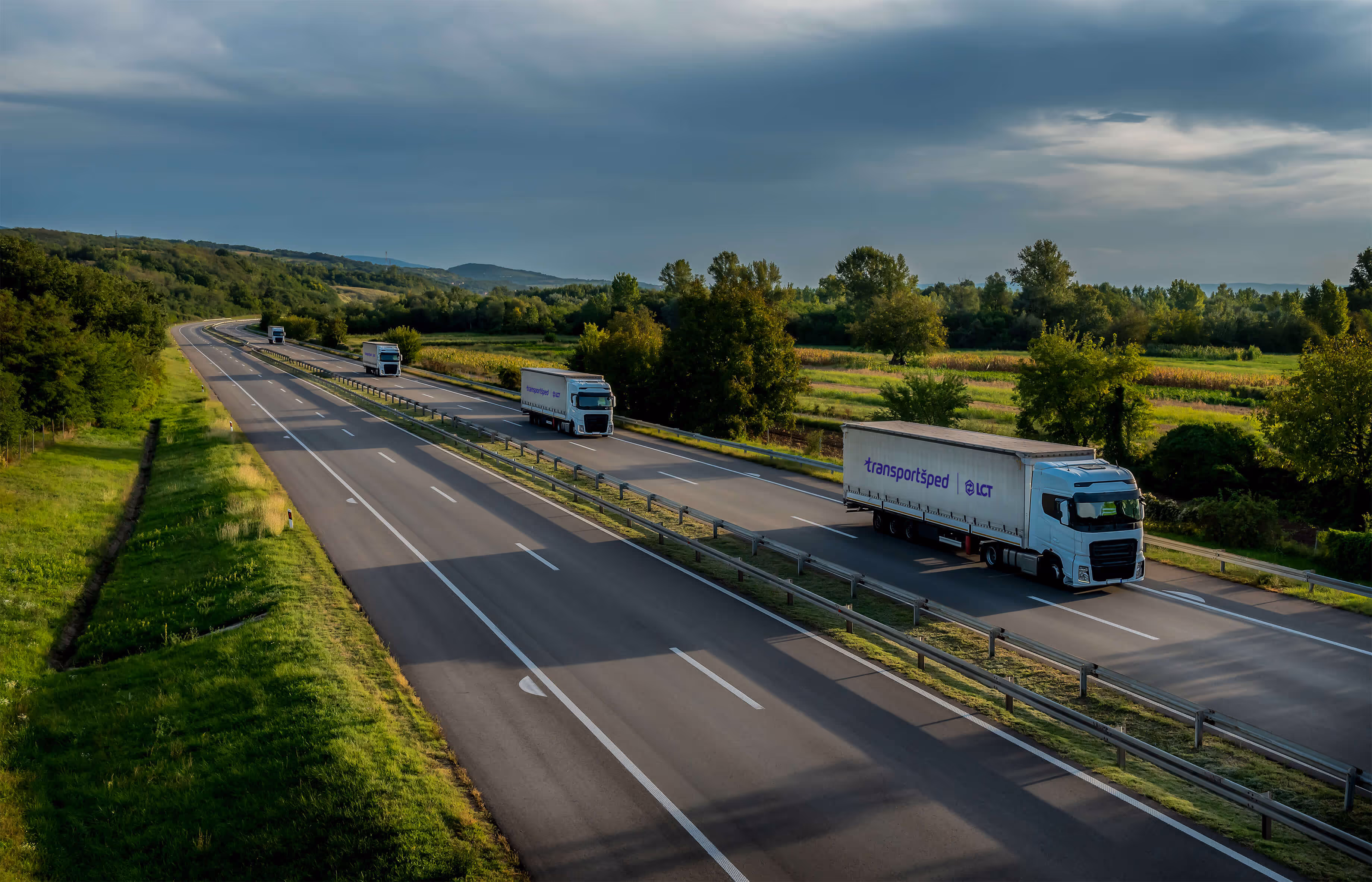 Three white transport trucks with 'transportsped LCT' logos driving on a divided highway surrounded by green fields and trees under a cloudy sky.
