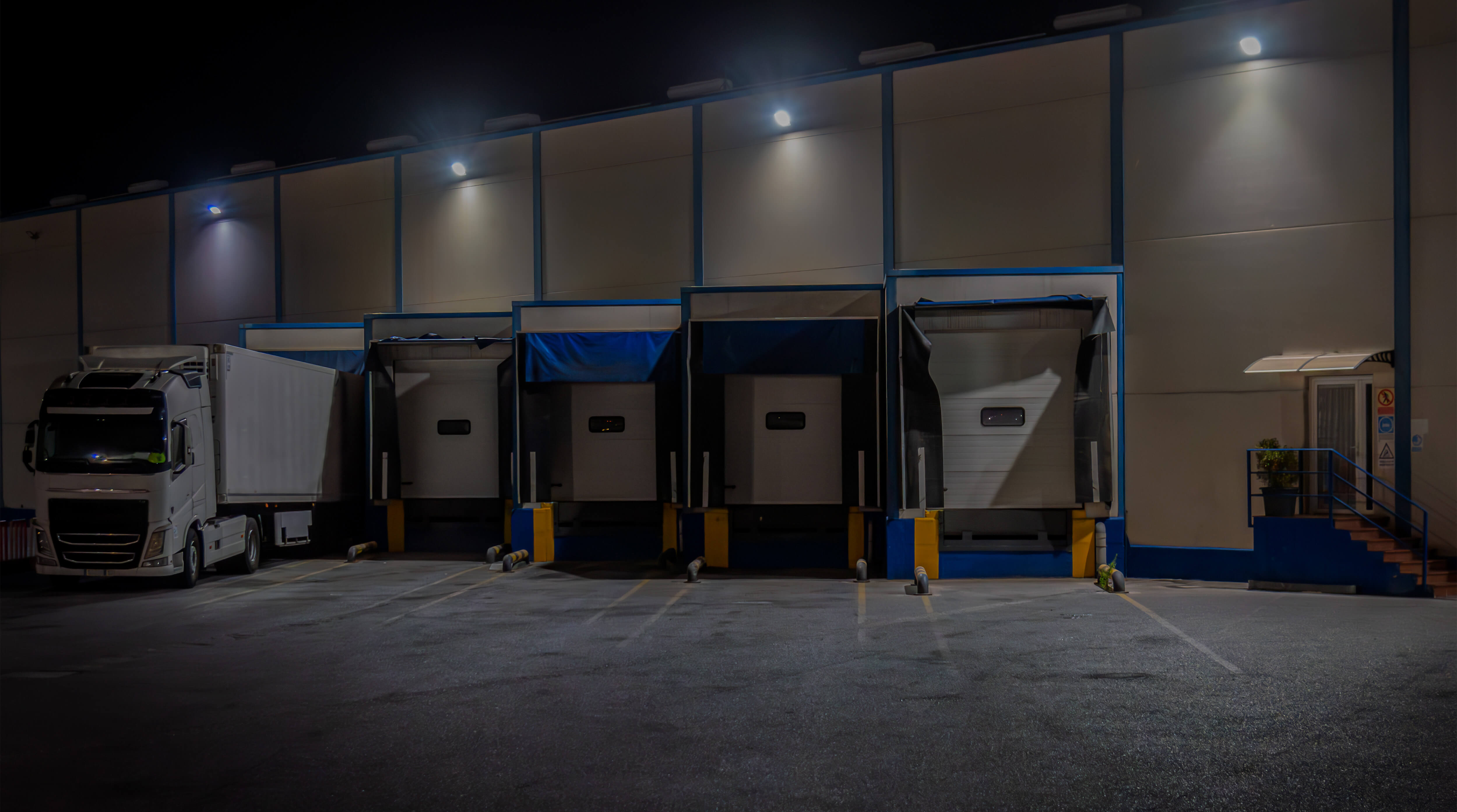 A large white semi-truck parked at a warehouse loading dock with five closed loading bay doors illuminated at night.