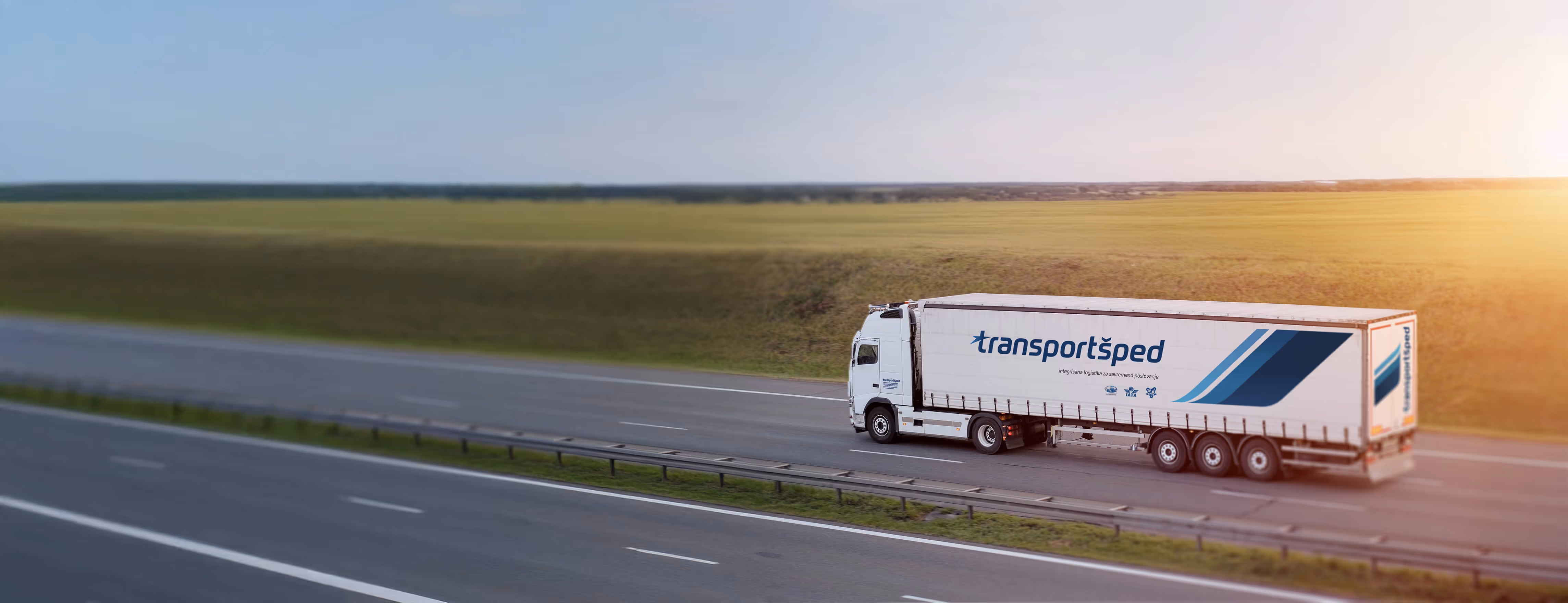 White semi-truck with Transportsped logo driving on a highway alongside green fields during sunset.