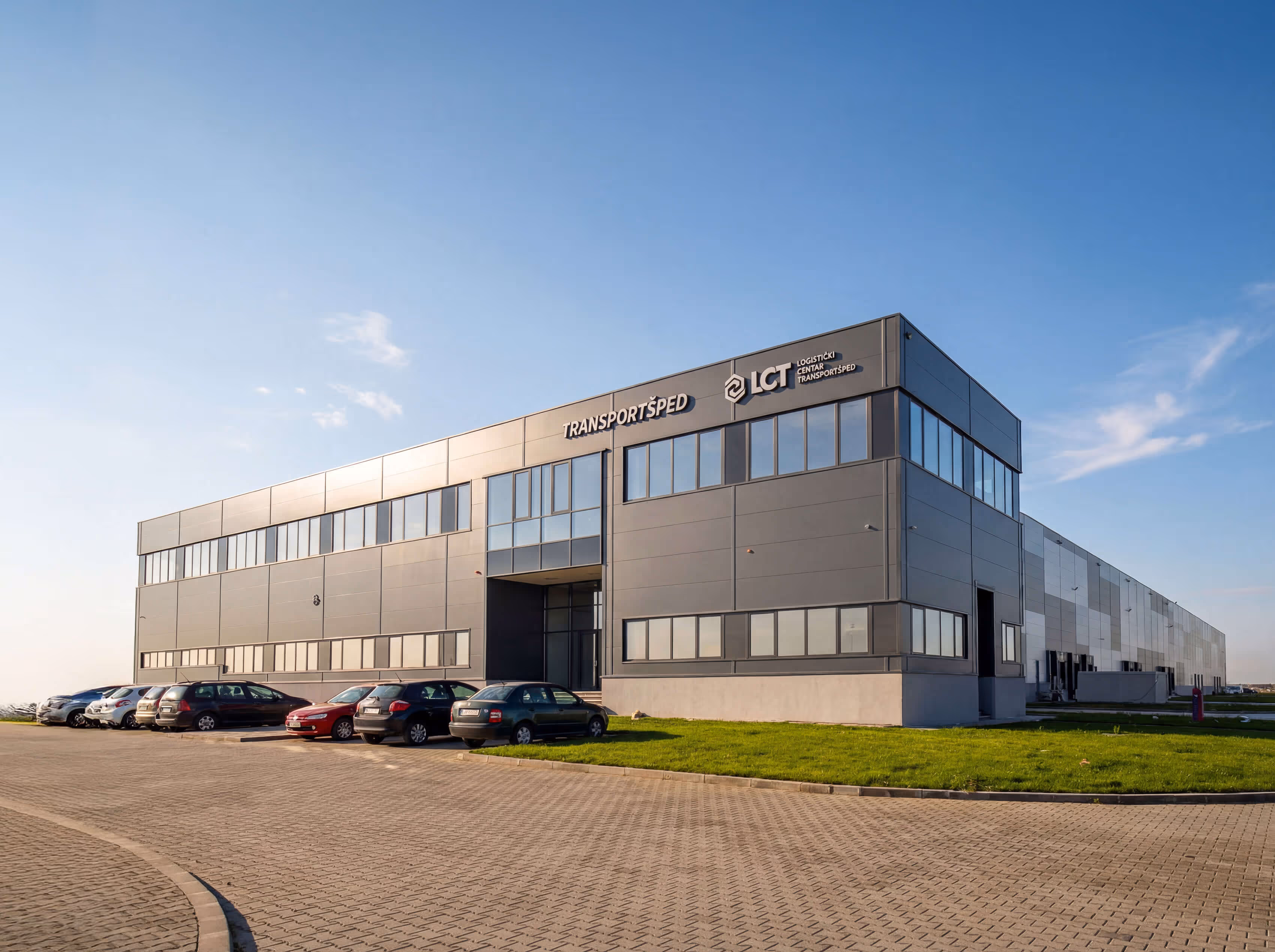 Modern industrial building with TRANSPORTŠPED and LCT logos, parked cars, and clear blue sky.