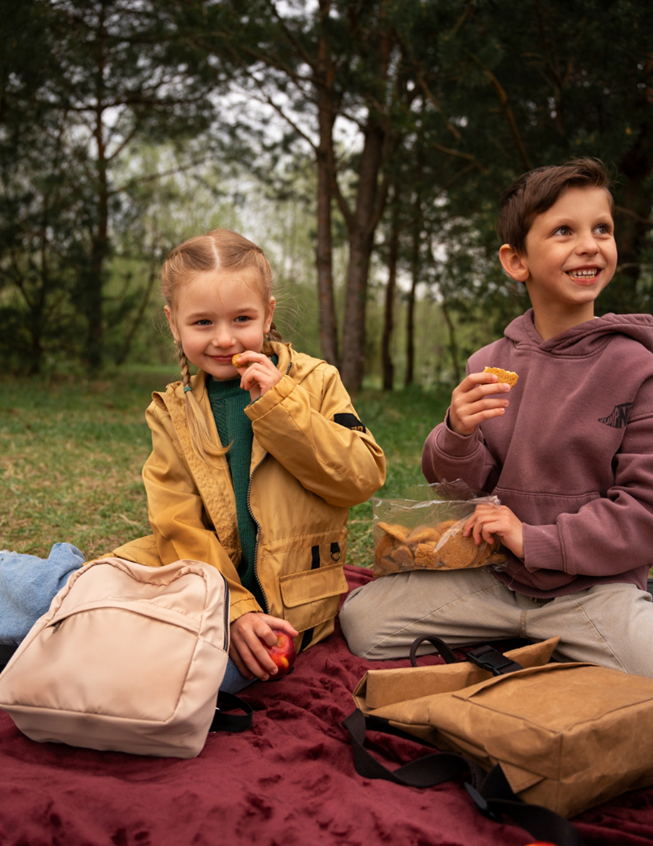 Two children sitting on a blanket outdoors, eating cookies and smiling during a picnic.