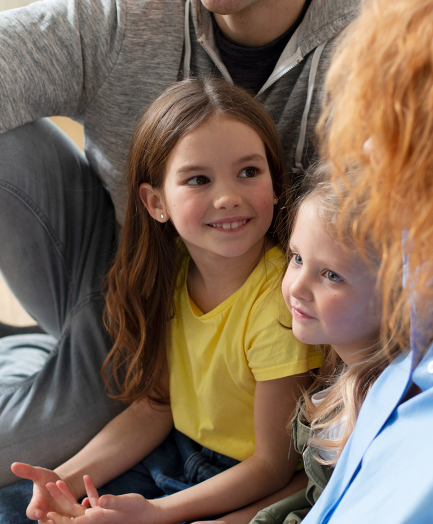 Two young girls sitting closely and smiling, engaged in a conversation with a partially visible adult.