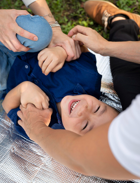 Child lying on a picnic blanket outdoors, smiling and being playfully held by multiple adults.
