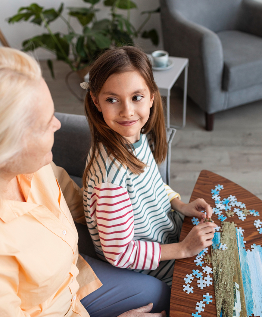 Young girl and elderly woman sitting together at a table, assembling a jigsaw puzzle with landscape image.