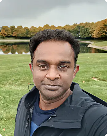 Man with short curly hair wearing a black jacket, standing in a grassy park with a lake and trees in the background under a cloudy sky.