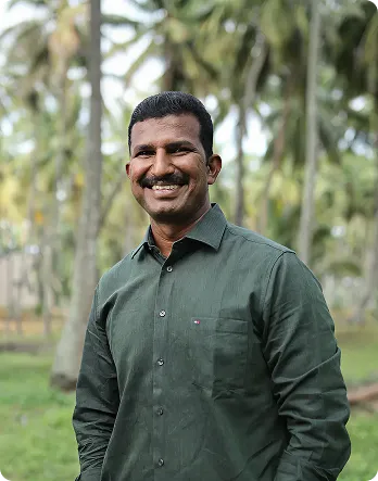 Smiling man in a green button-up shirt standing outdoors with trees in the background.