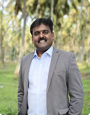 Smiling man in a light blue shirt and gray suit jacket standing outdoors with blurred trees in the background.