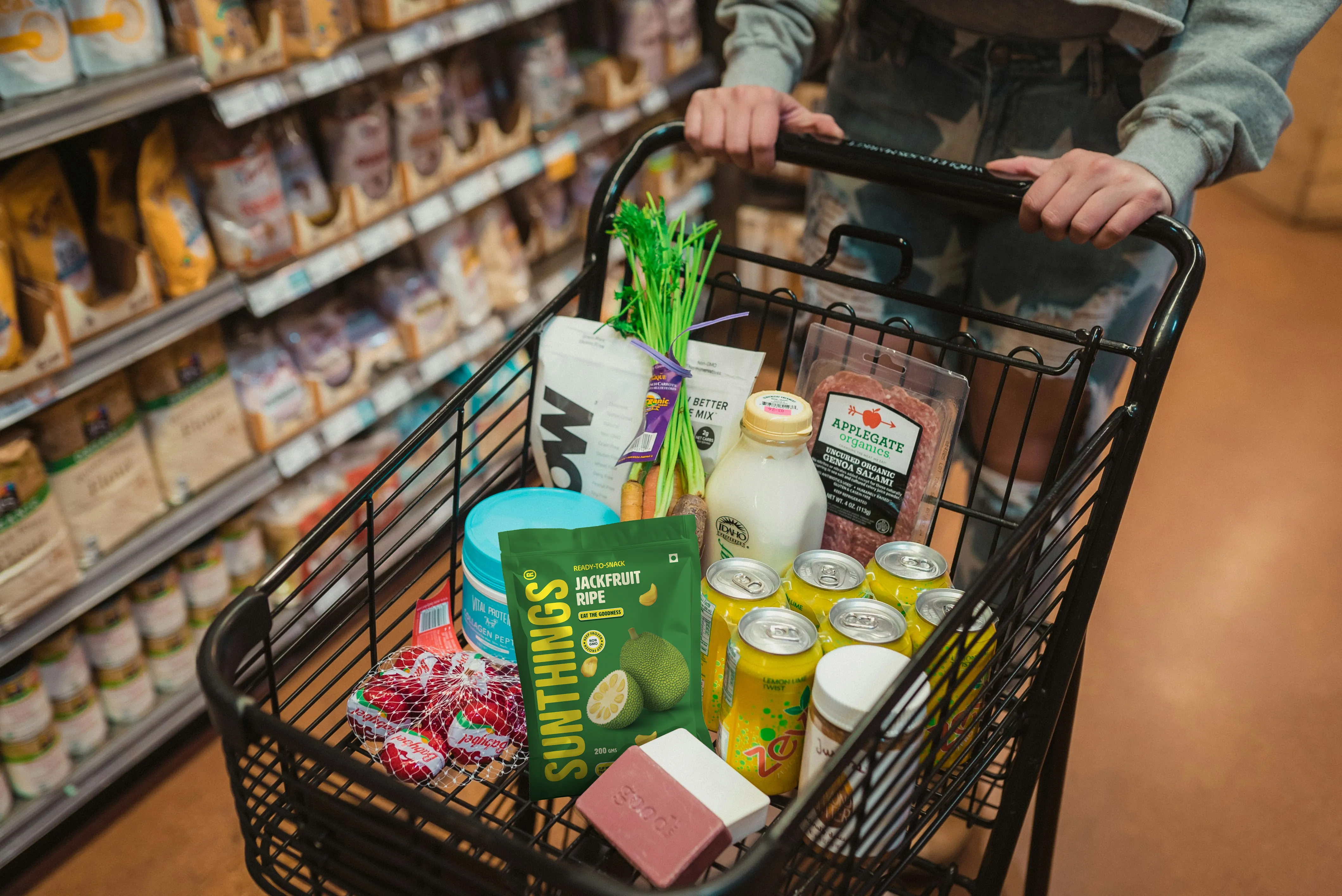 Shopping cart filled with various groceries including canned beverages, hand-grated coconut, a bottle of milk, organic salami, fresh vegetables, and packaged snacks.