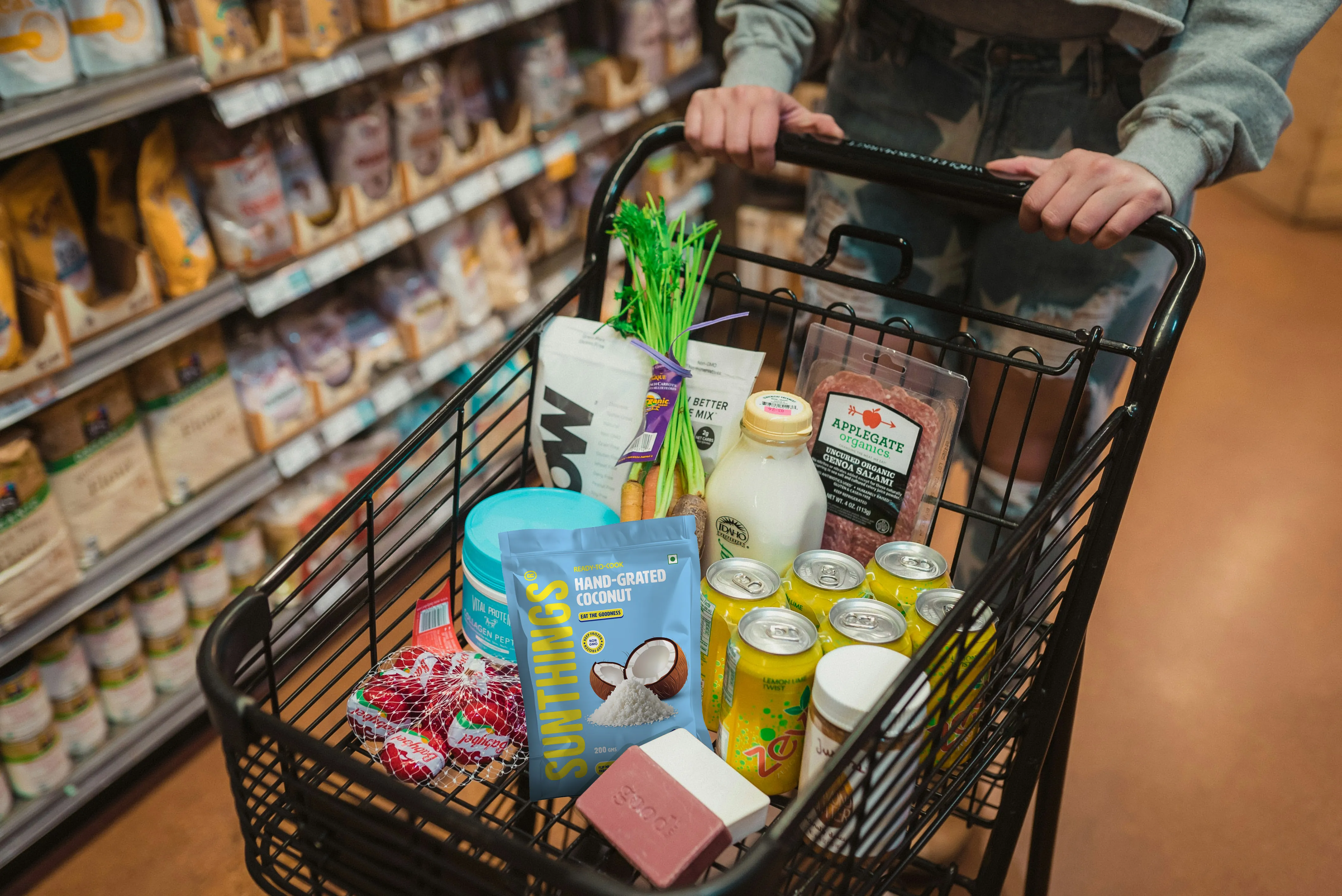 Grocery cart filled with various organic and health food items including a bag of hand-grated coconut, several yellow soda cans, packaged salami, and fresh produce.
