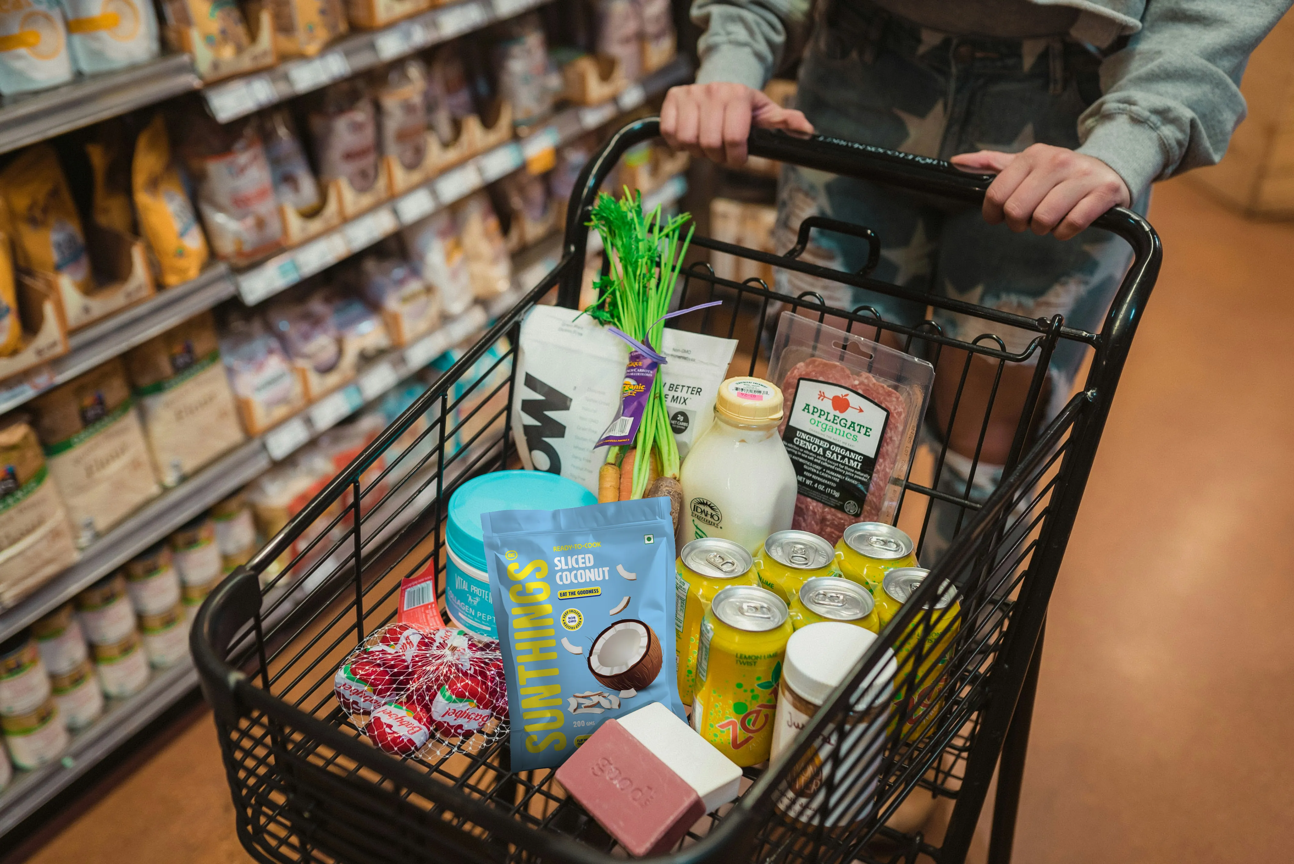 Shopping cart filled with groceries including sliced coconut, celery, canned drinks, milk, salami, cherry tomatoes, and a soap bar, pushed by a person in a gray sweatshirt and ripped jeans inside a supermarket aisle.