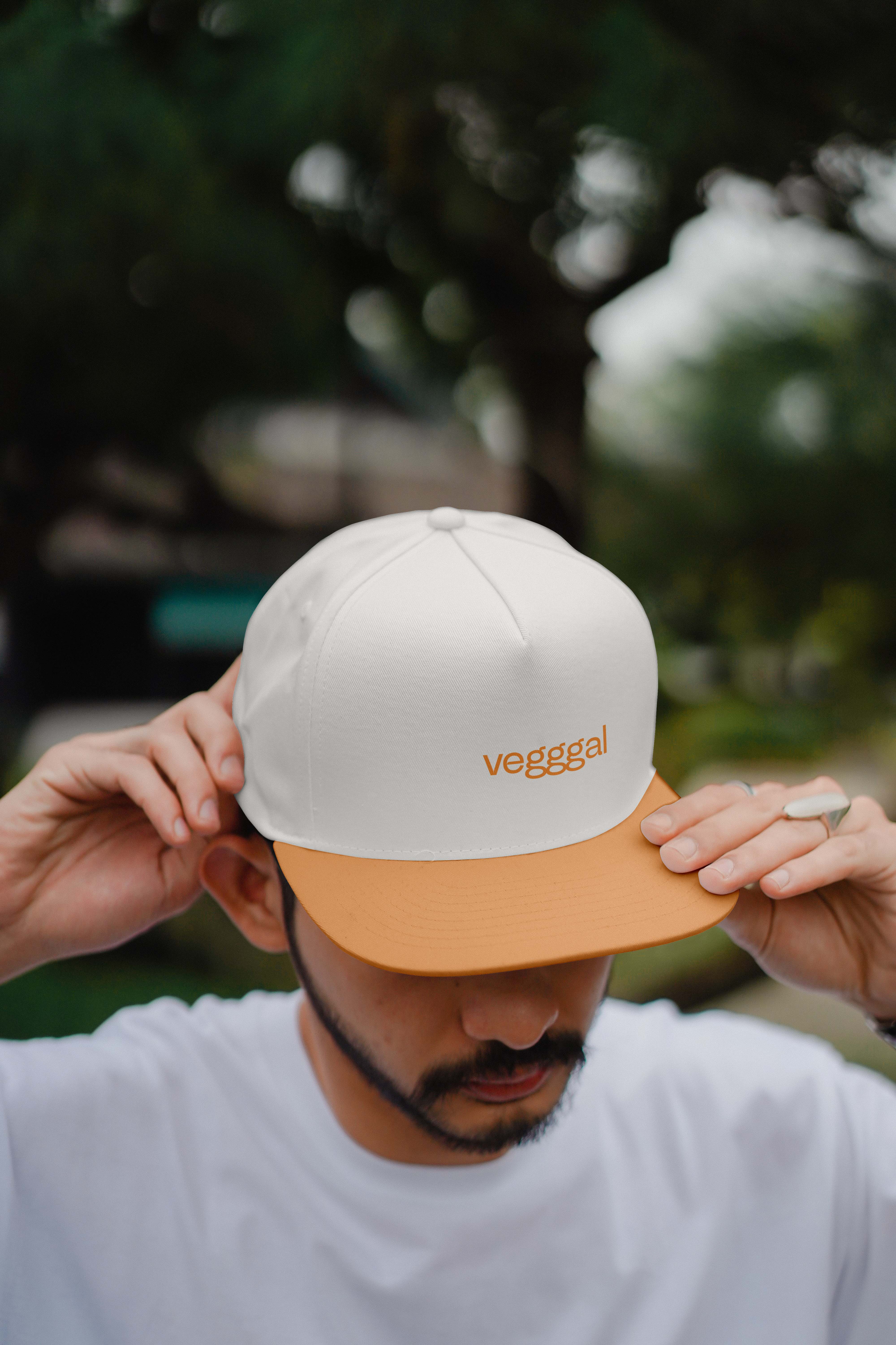 Mann med hvit og oransje caps med teksten 'vegggal' og hvit T-skjorte, justerer capsen utendørs.