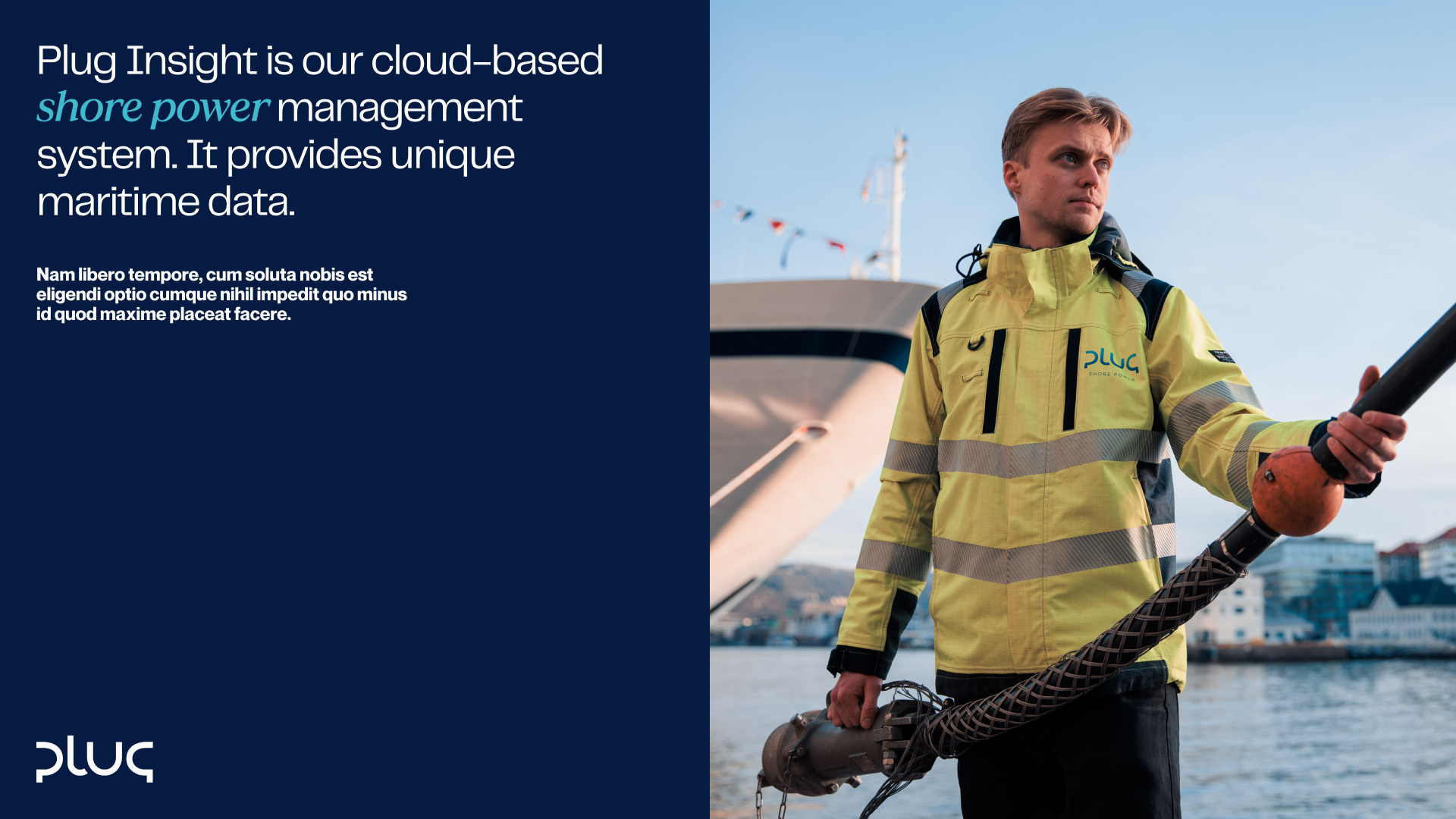 Man in yellow high-visibility jacket holding a thick cable by the water with a ship and buildings in the background.