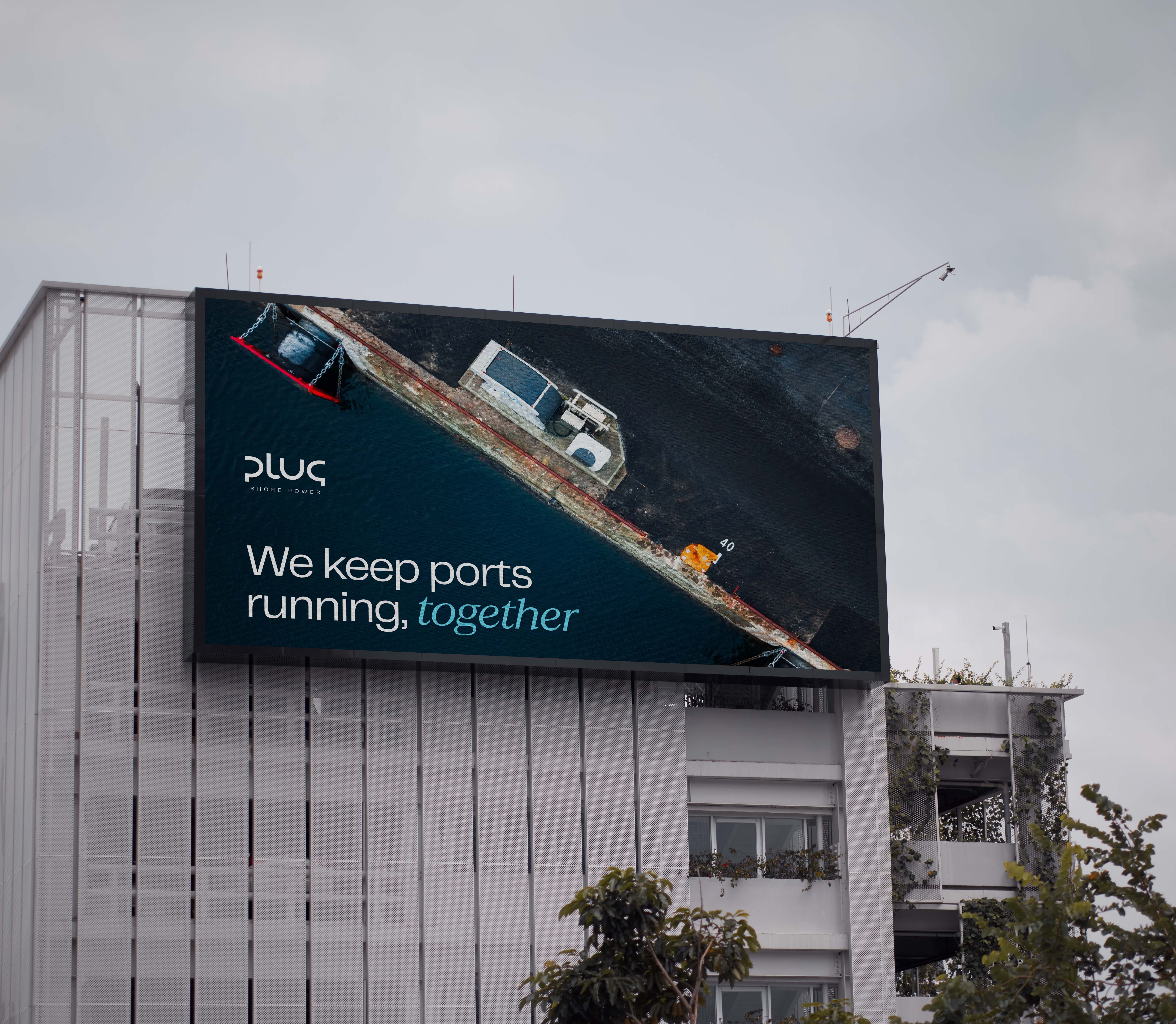 Billboard on a building showing an aerial view of a port dock with the text 'We keep ports running, together' and the Plug Shore Power logo.