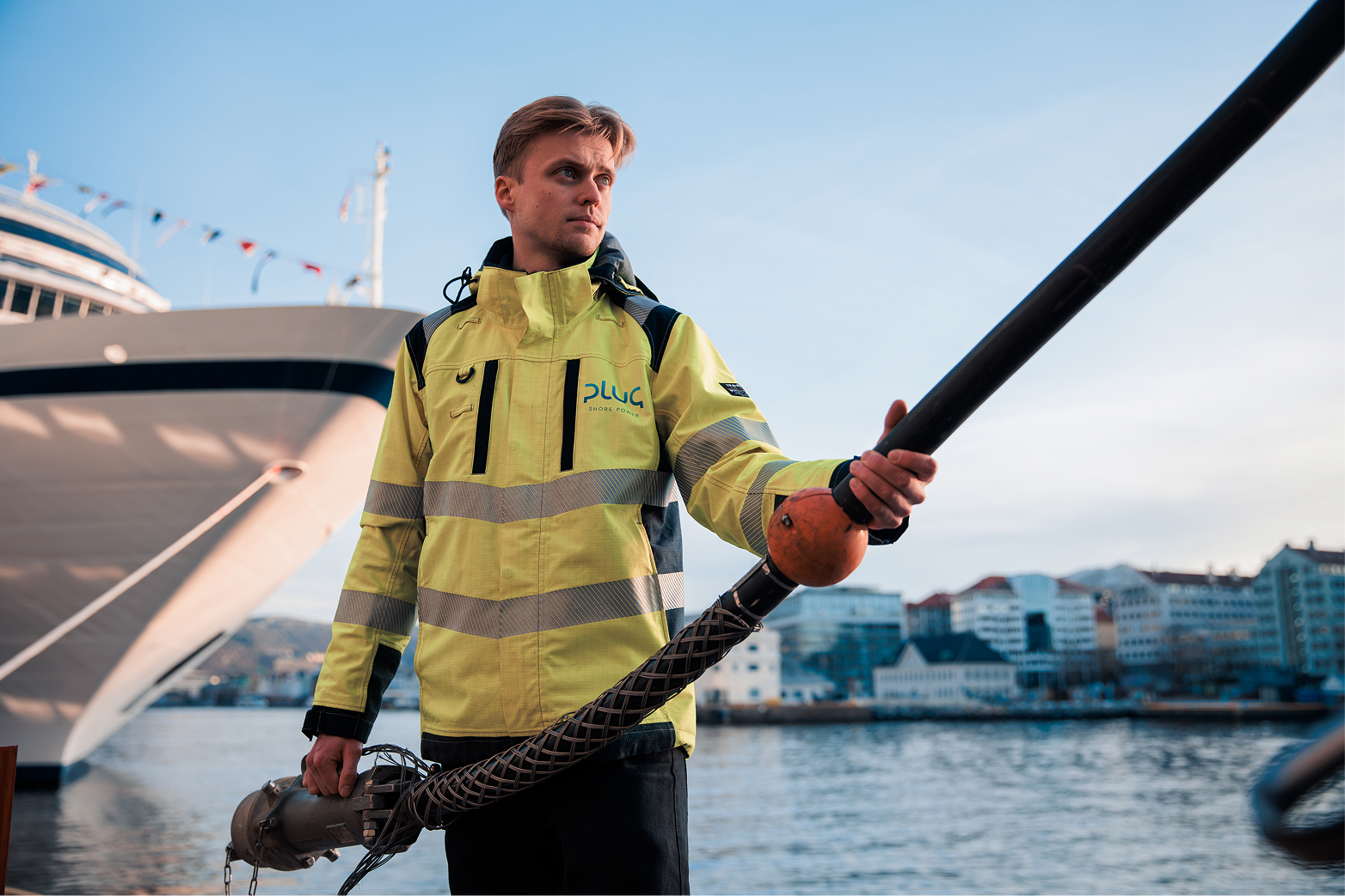 Man in a yellow reflective jacket holding a large black marine cable near a docked ship with city buildings in the background.