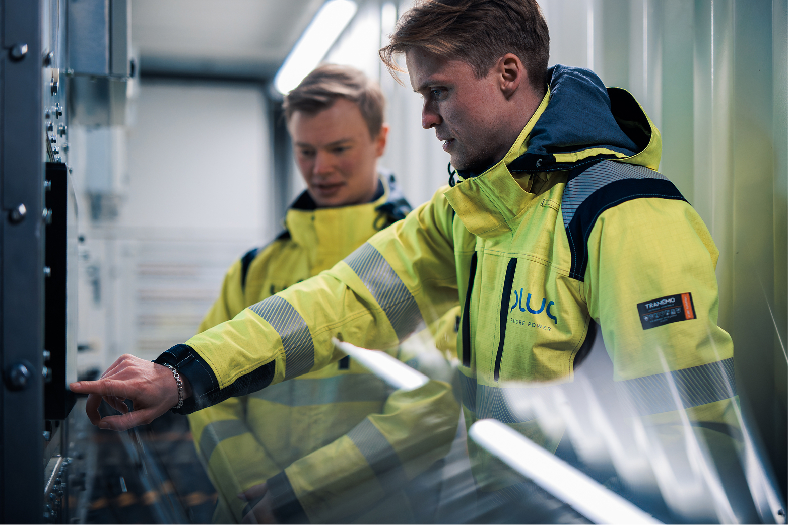Two men in yellow high-visibility jackets operating electrical equipment indoors.
