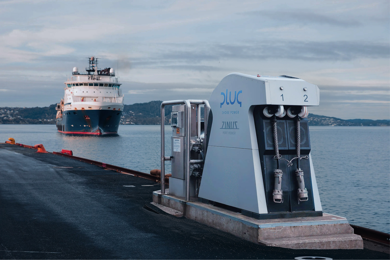Dockside shore power unit labeled 'Plug Shore Power Zinus Port Power' with a ship approaching in the background on calm water under a cloudy sky.