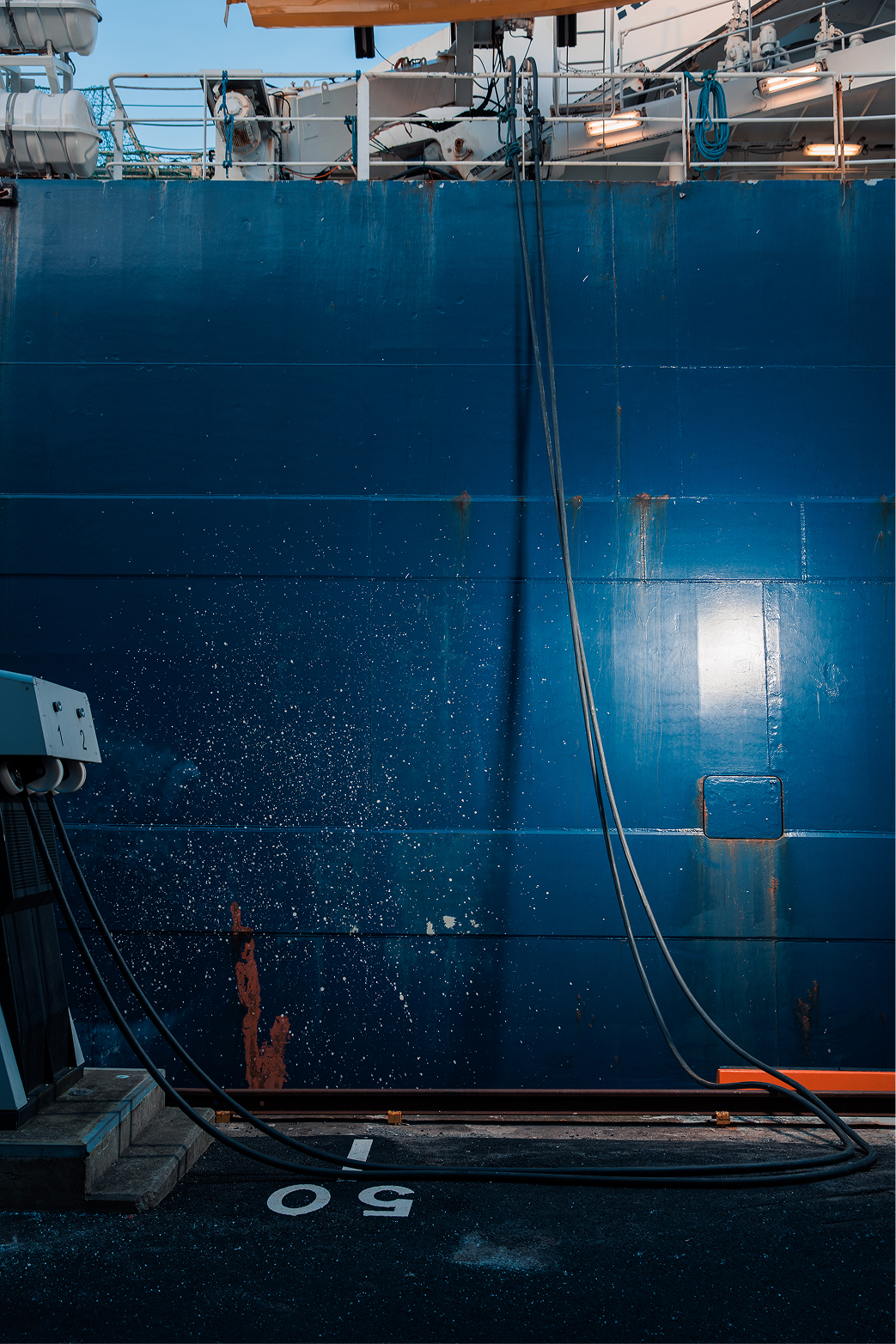 Fuel hoses connected to a large blue ship hull with water splashing nearby at a dock.
