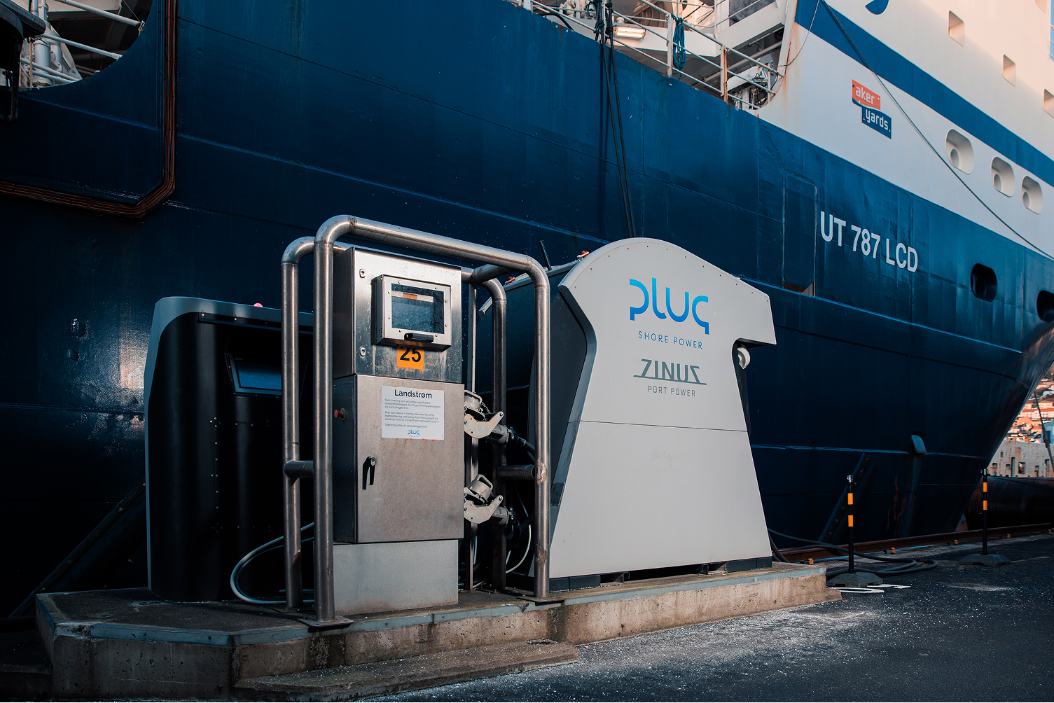 Electric shore power supply unit labeled Plug and Zinus on a dock beside a large blue and white ship marked UT 787 LCD.