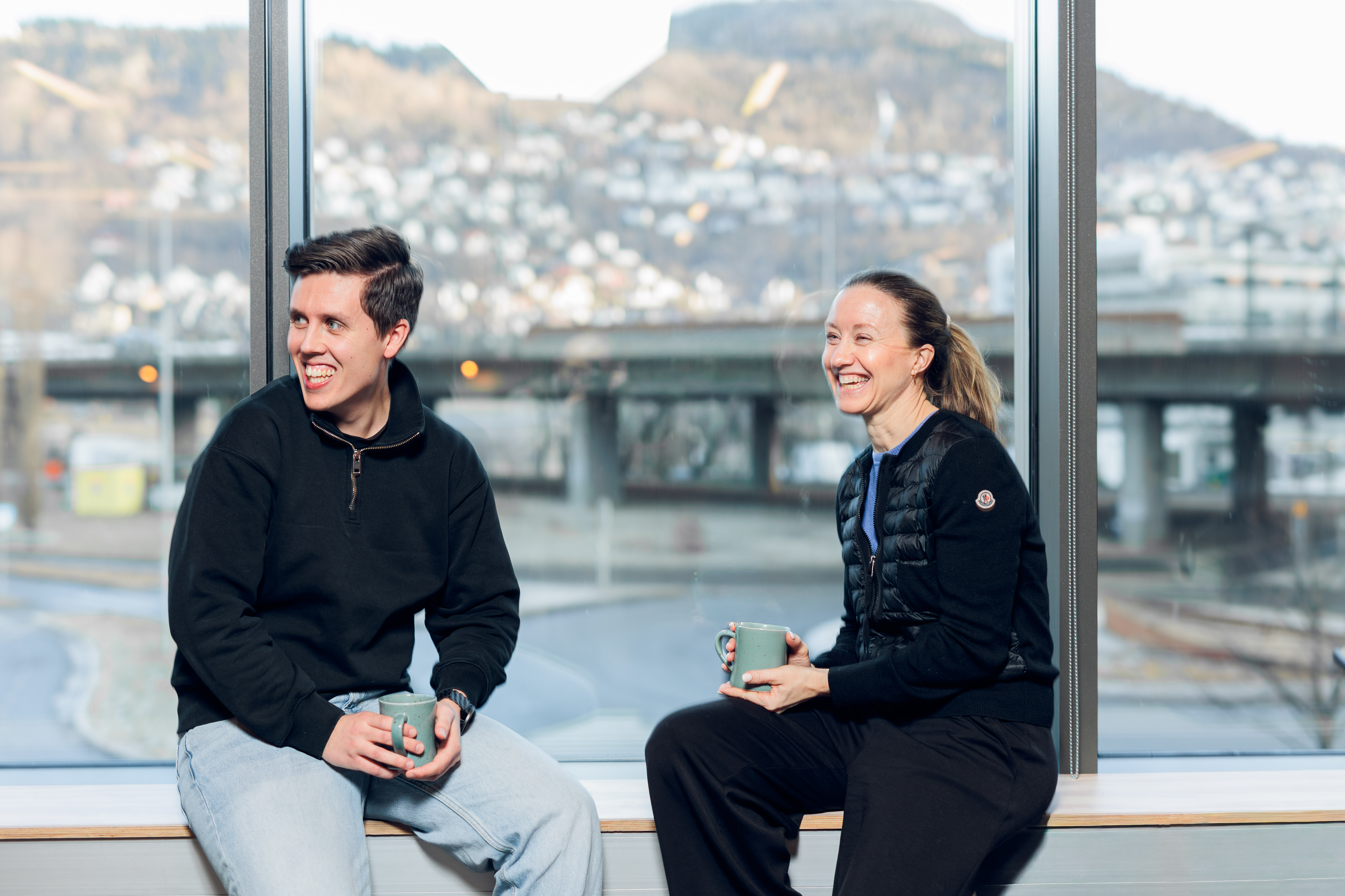 Two people sitting on a bench by a large window, smiling and holding green mugs, with a blurred cityscape outside.