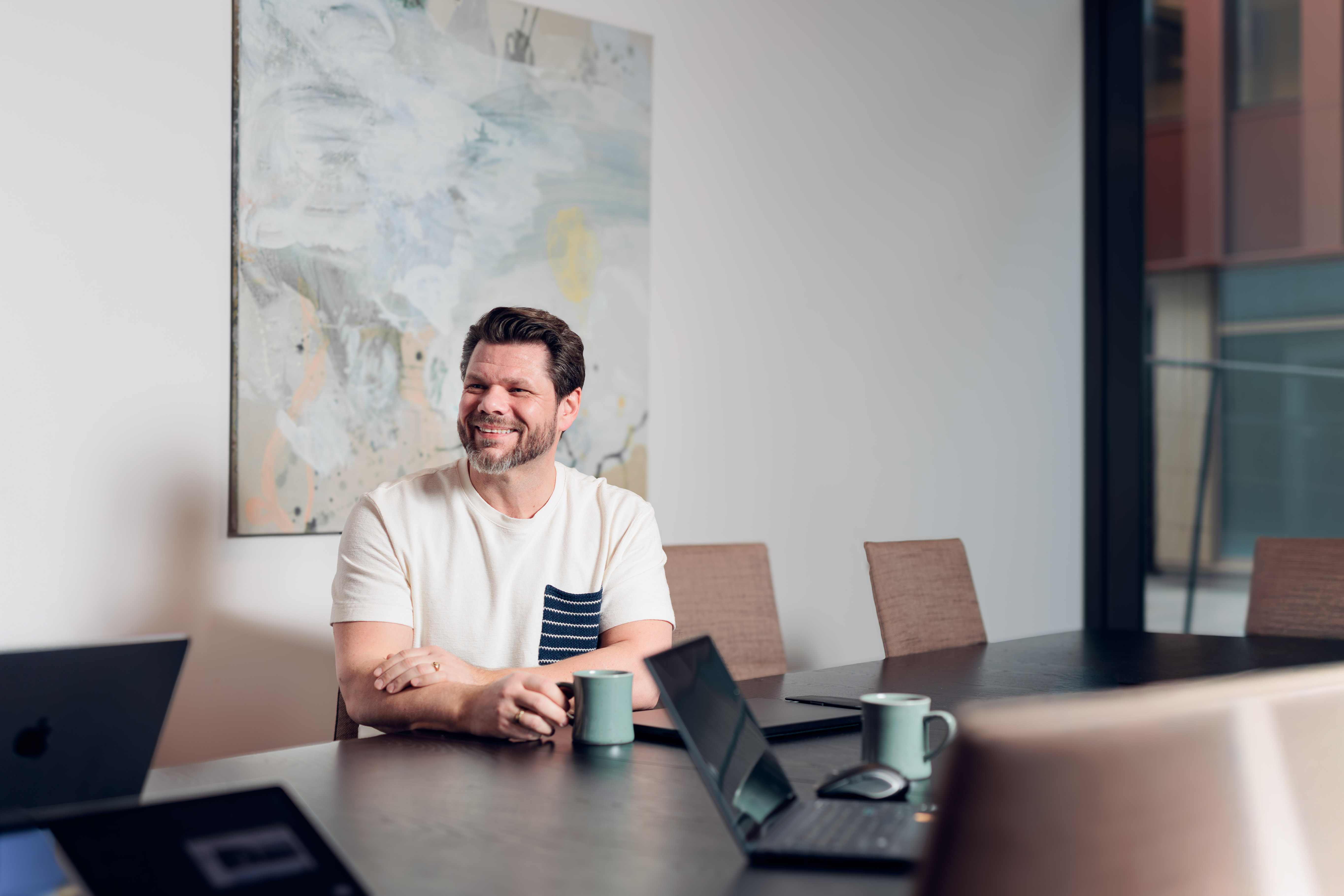 Smiling man in white shirt sitting at a conference table with laptops and mugs.
