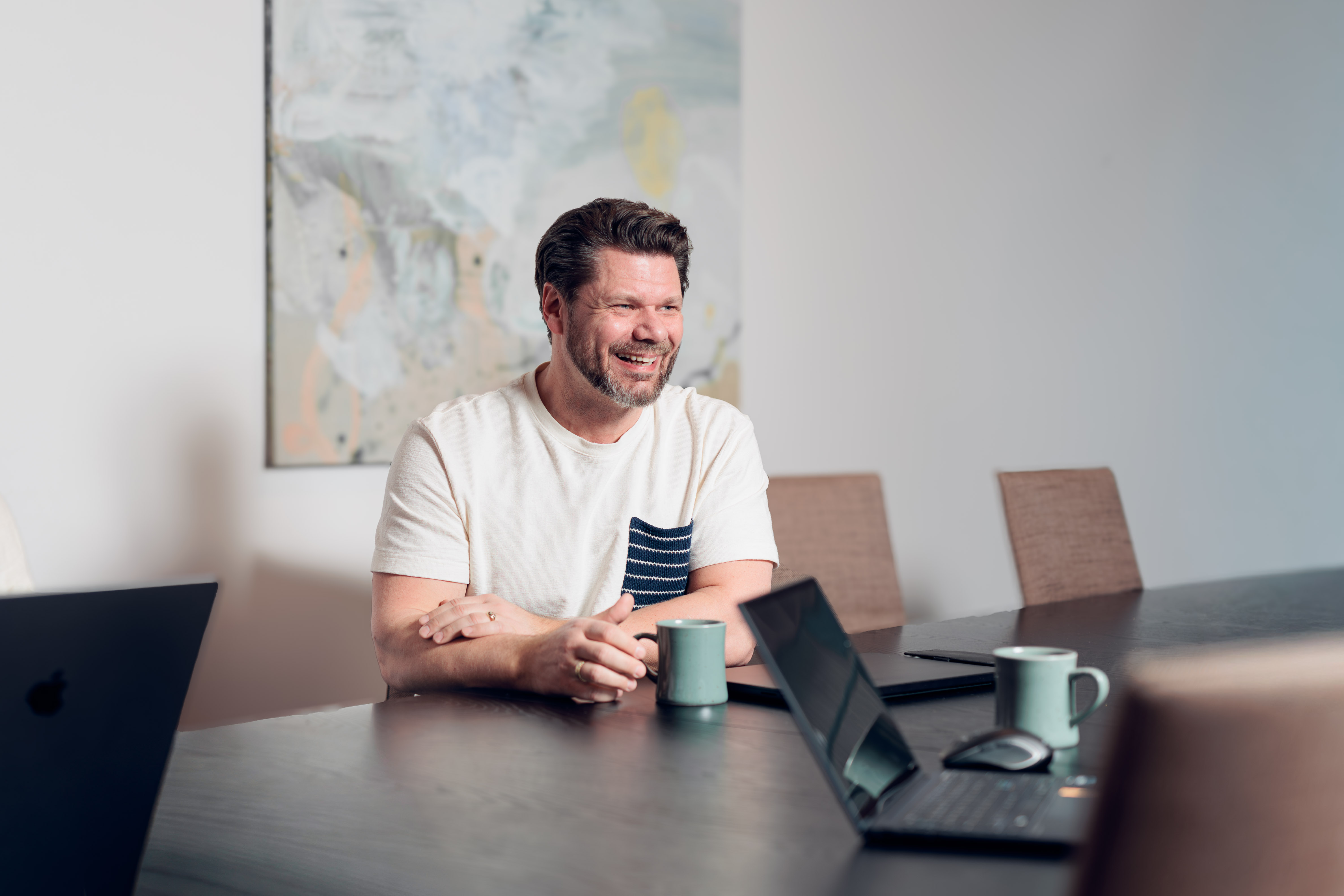 Smiling man wearing a white shirt with a striped pocket, sitting at a conference table with laptops and coffee mugs.