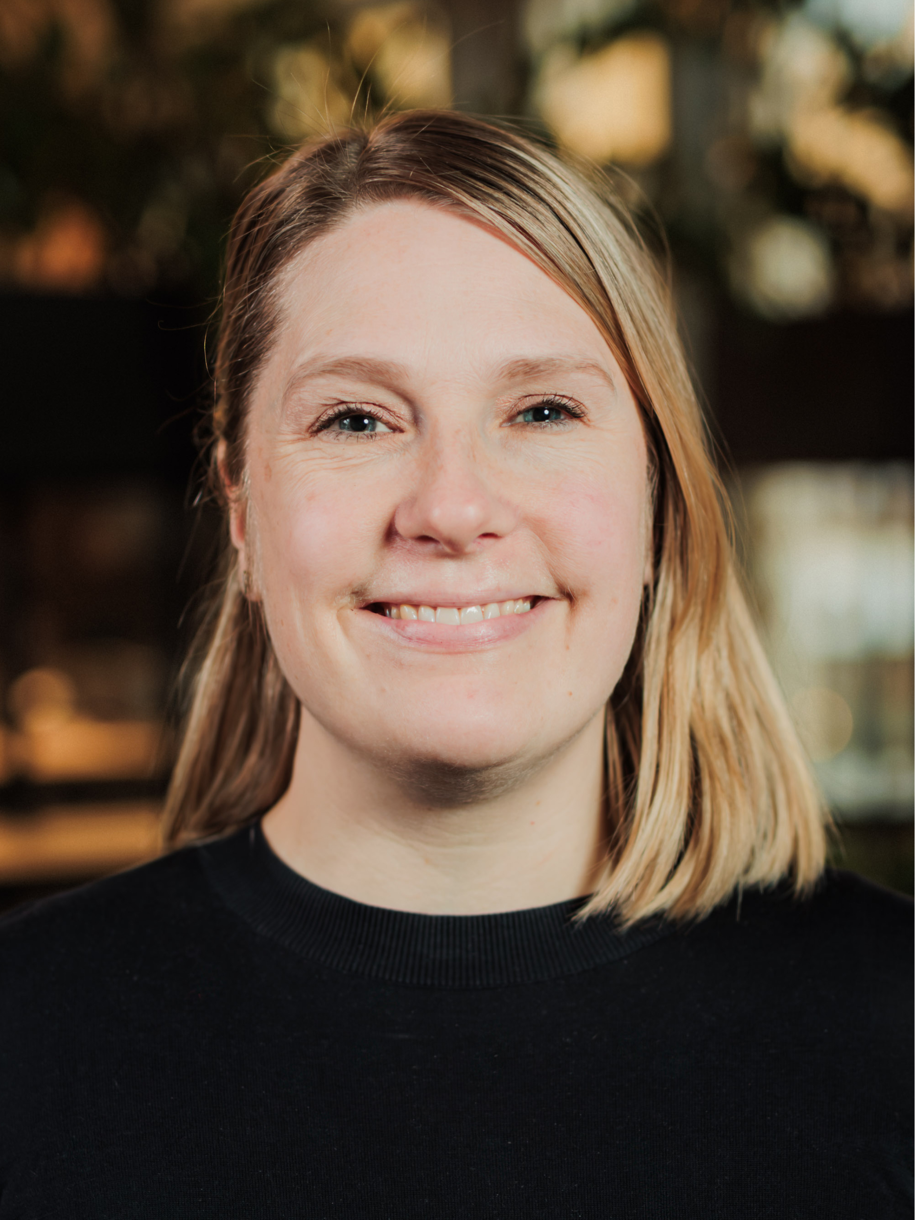 Smiling woman with shoulder-length blonde hair wearing a black top, with a blurred warm background.