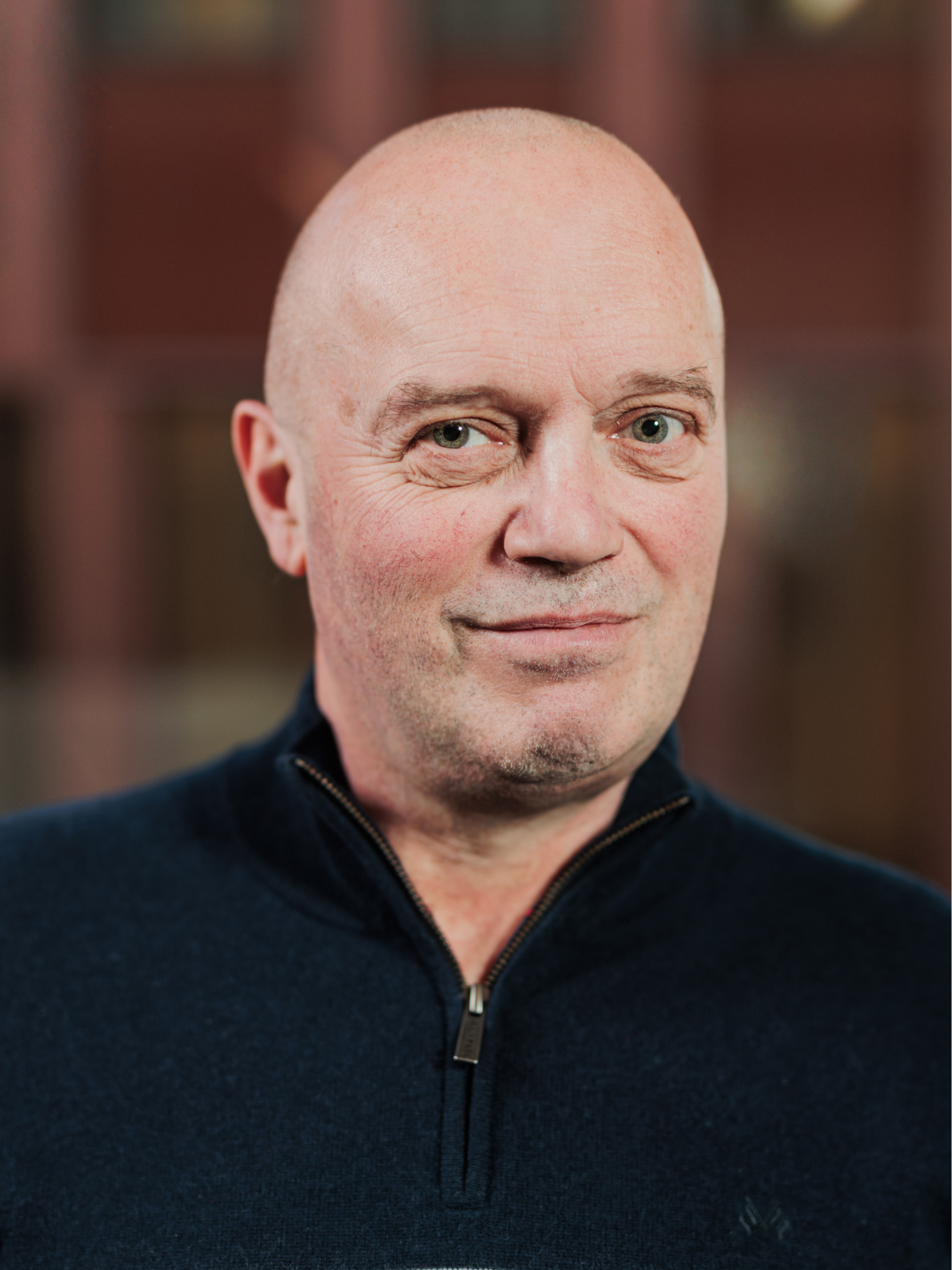 Portrait of a bald man with green eyes wearing a dark blue zip-up sweater, smiling slightly against a blurred background.