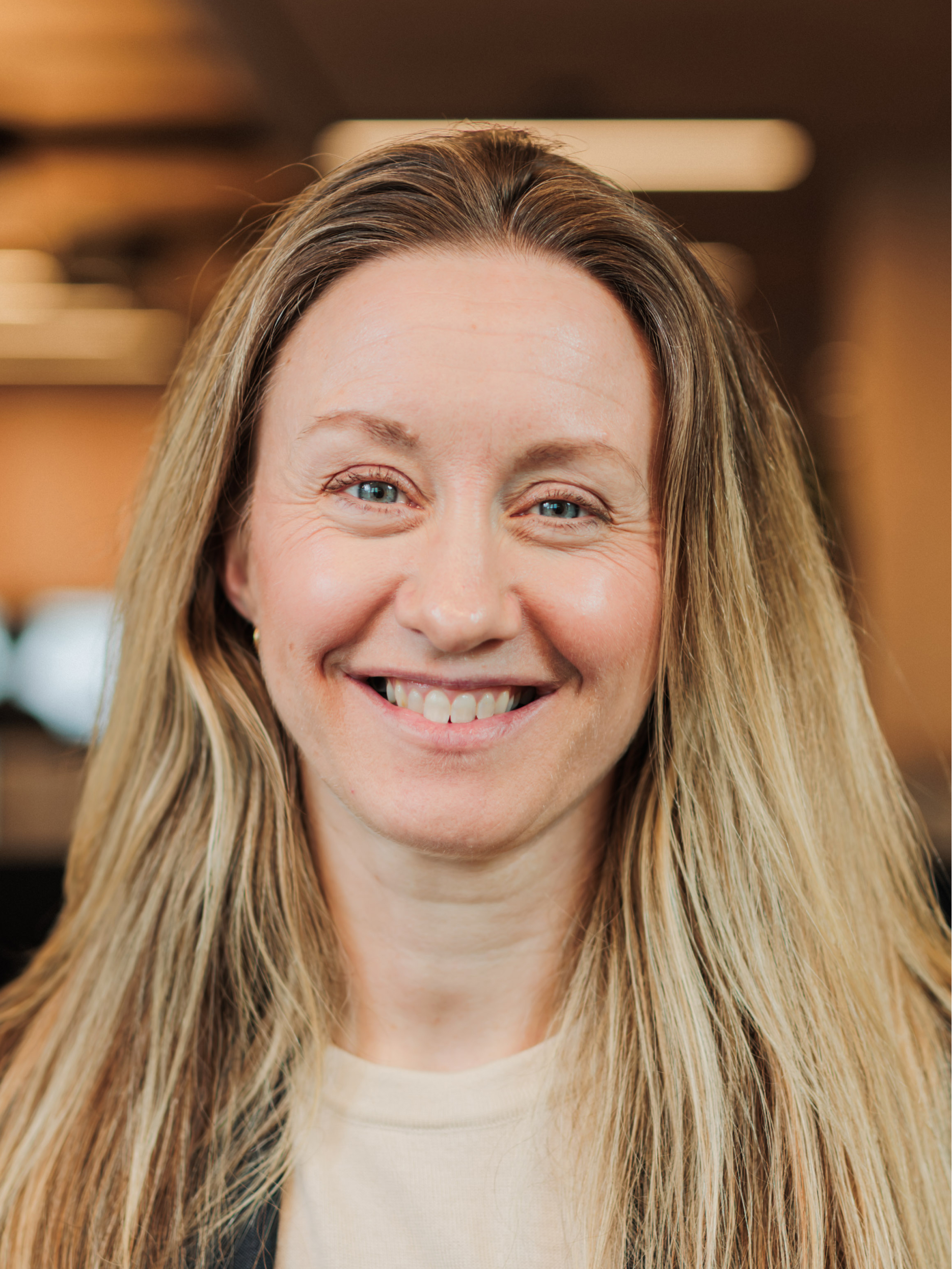 Smiling woman with long blonde hair and blue eyes, wearing a light top and dark jacket in an indoor setting.