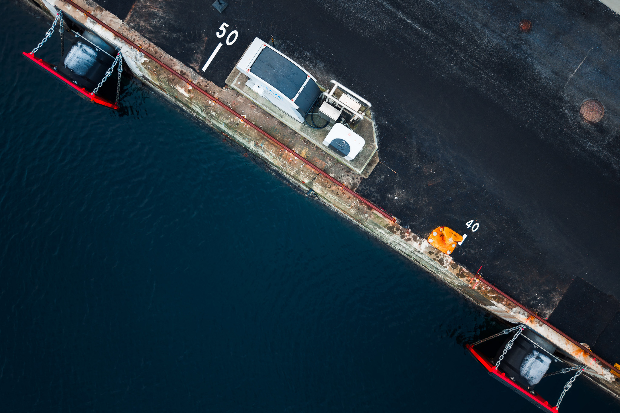 Overhead view of a weathered concrete dock with safety barriers, an orange cone, and calm dark blue water.