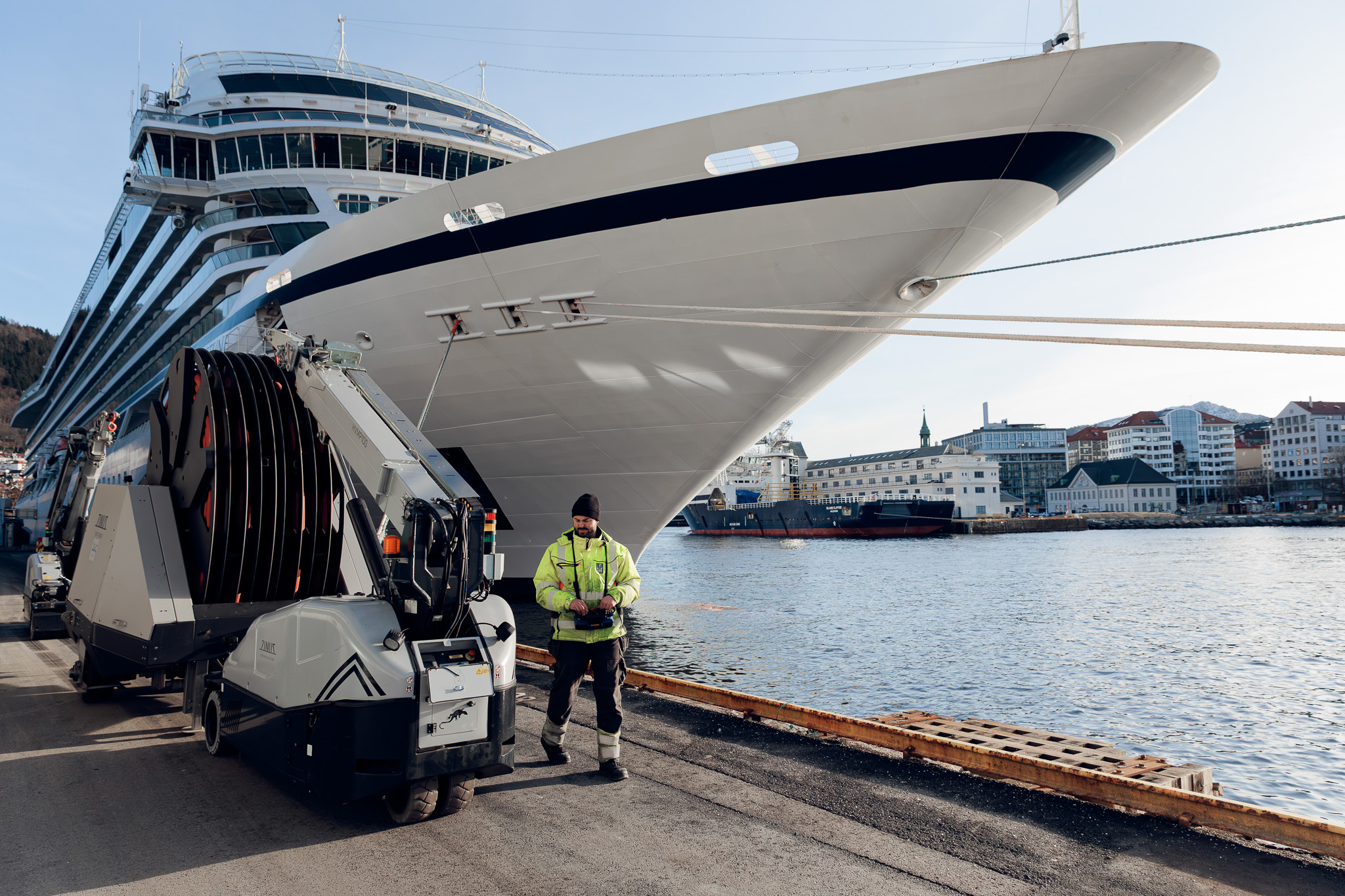 Worker in high-visibility jacket operating machinery on dock next to large cruise ship moored by waterfront buildings.